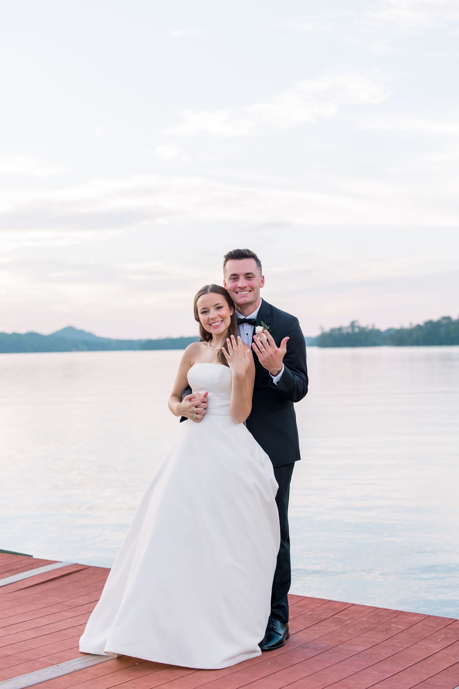 Newlywed couple showing wedding rings on a red dock by a lake at sunset, bride in white gown, groom in dark tuxedo.