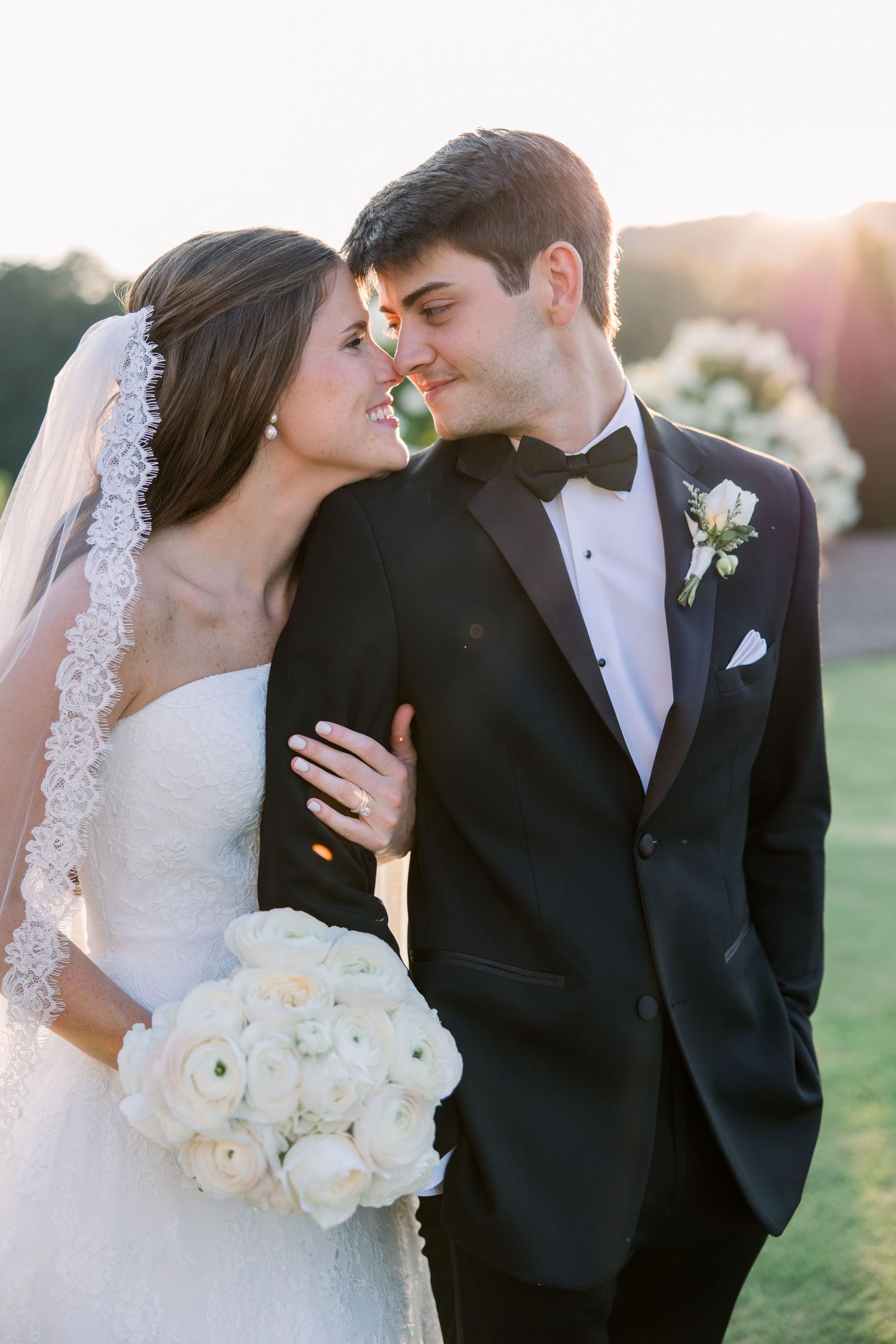 Bride and groom share a romantic moment at golden hour, she holds white ranunculus bouquet wearing lace veil.