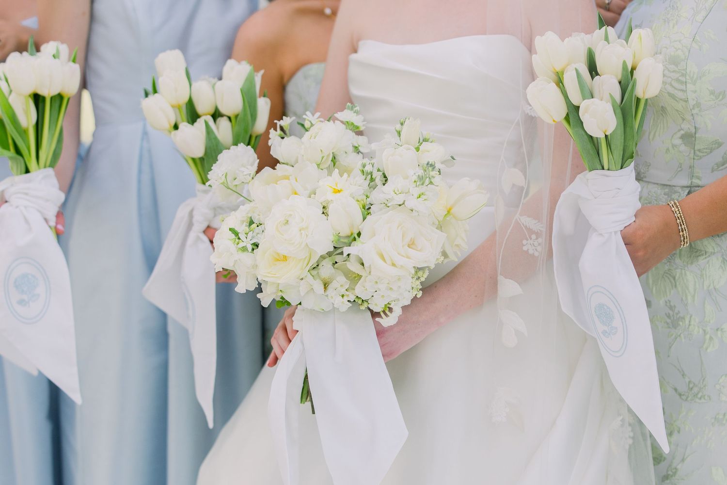 Bride and bridesmaids holding white tulip bouquets with satin ribbons, wearing white and light blue dresses.