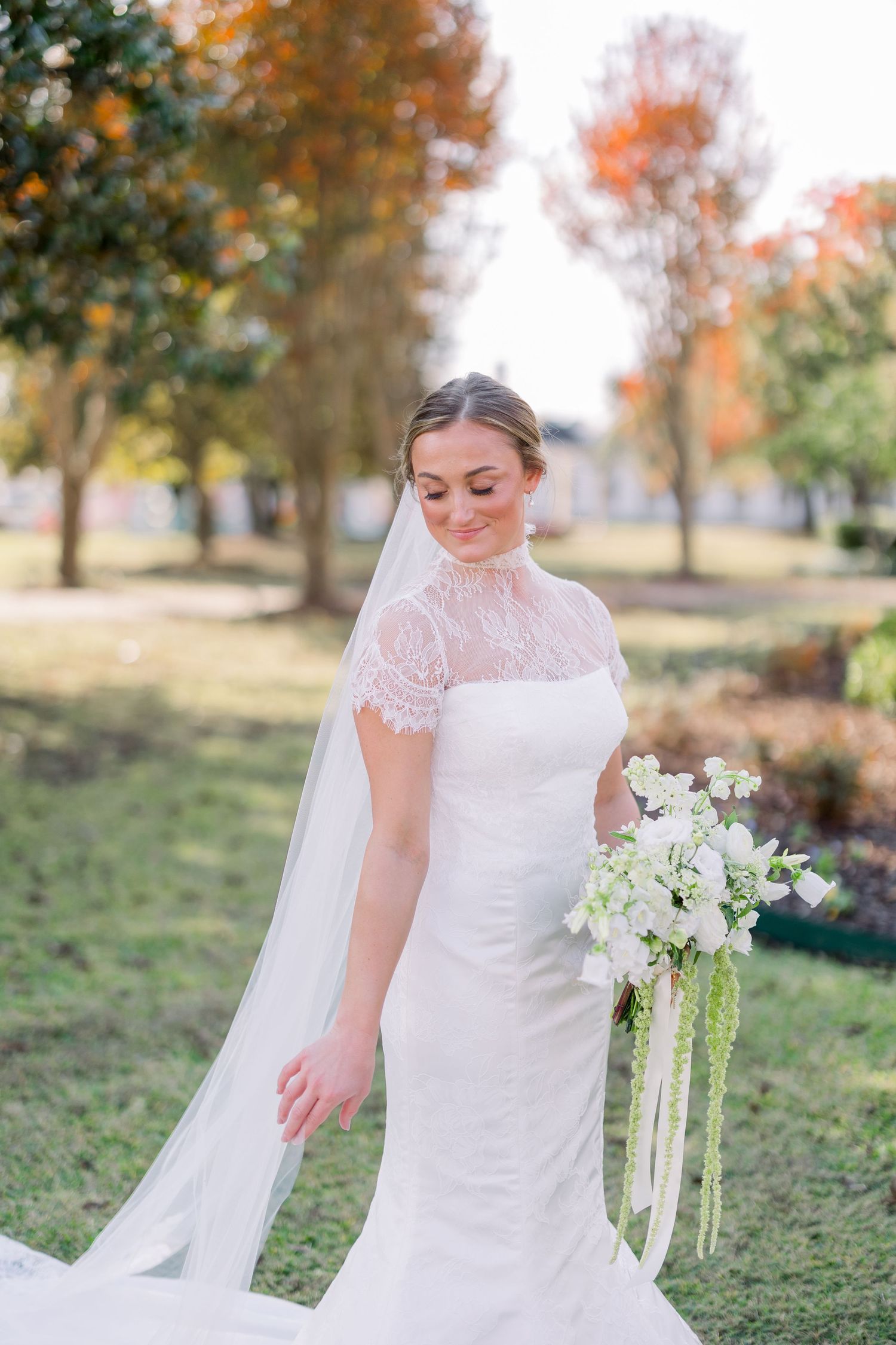 A bride in a lace-detailed wedding gown holds a white bouquet in an autumn outdoor setting.