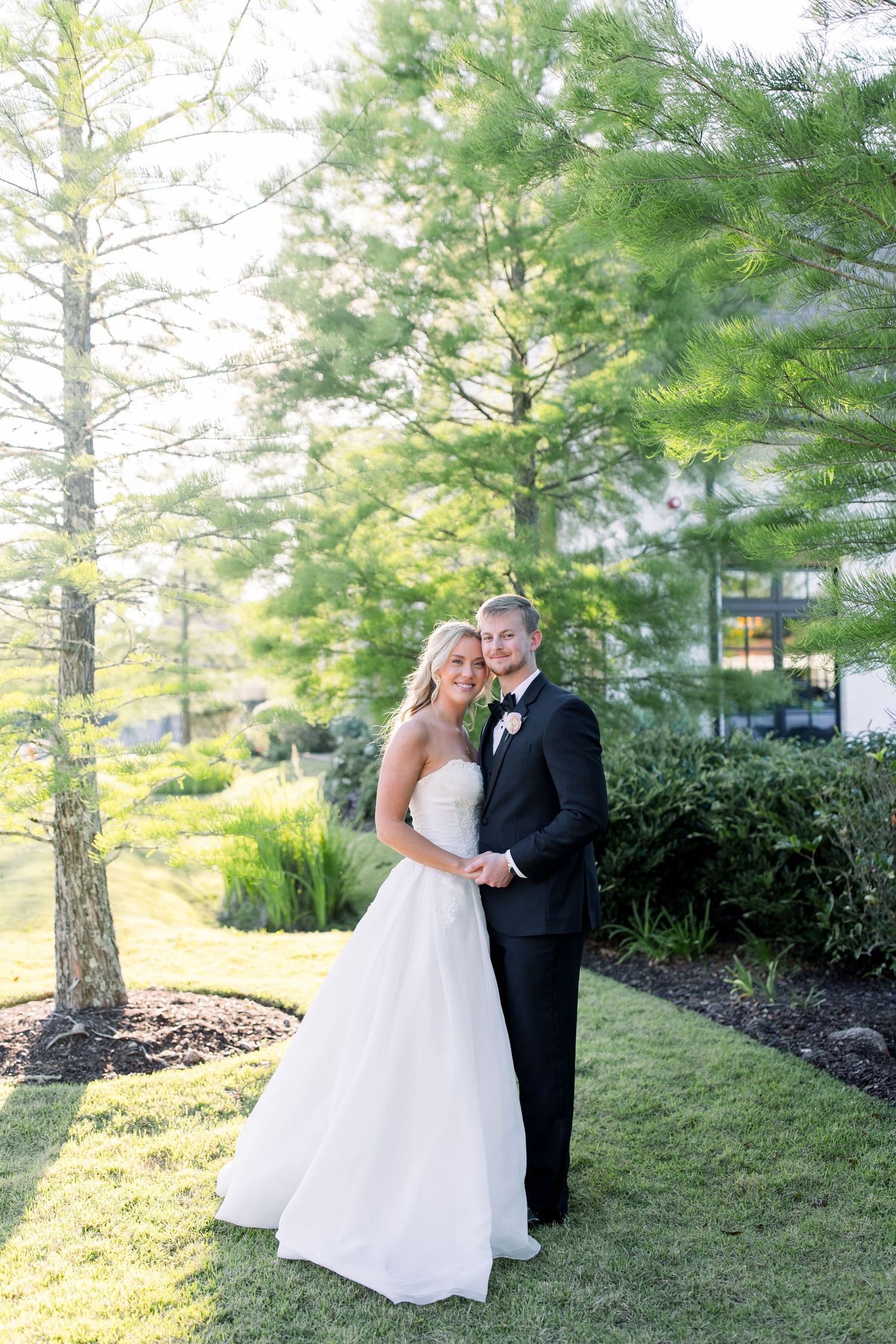 A bride and groom pose together outdoors on a lush green lawn surrounded by tall trees on their wedding day.