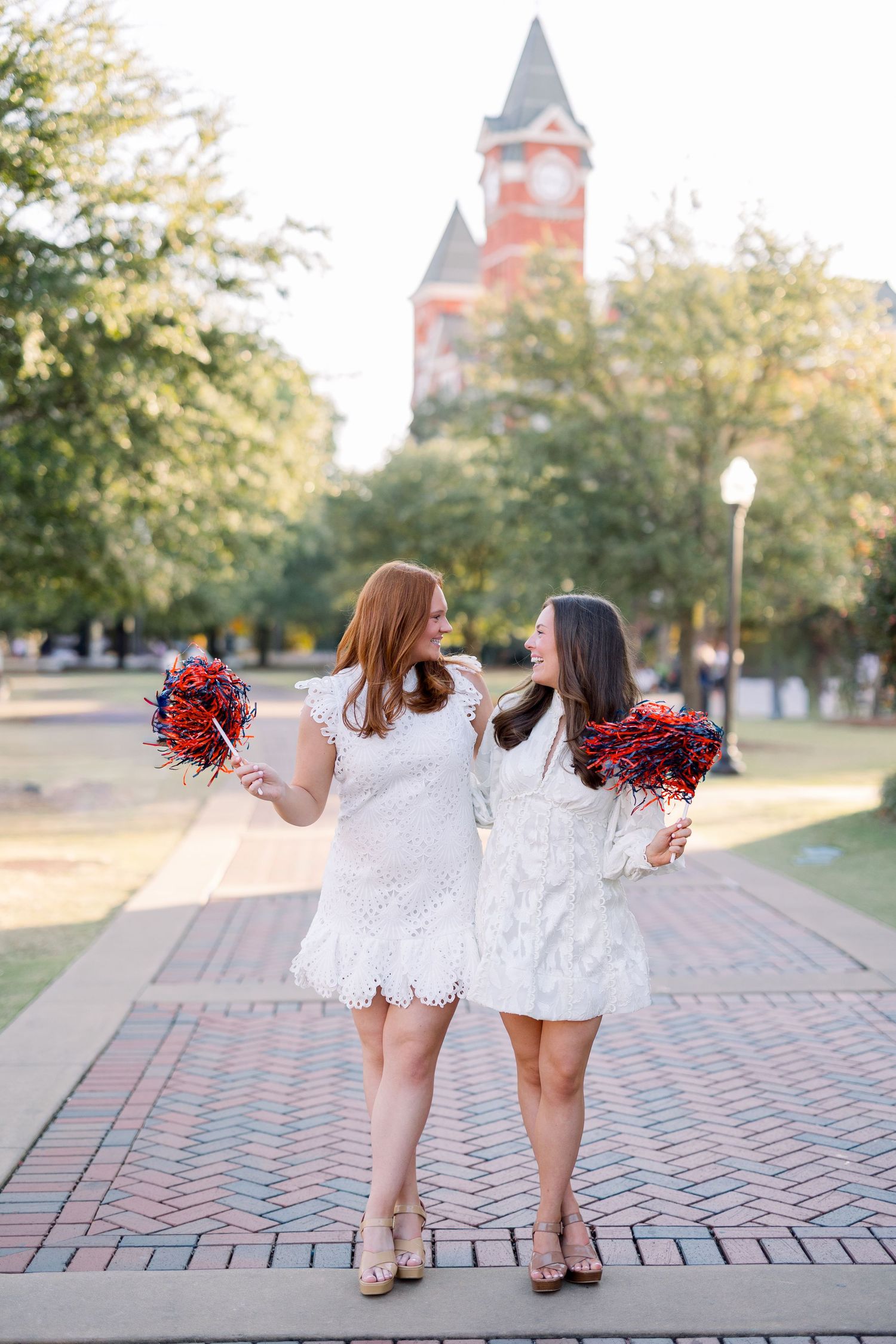 Two cheerleaders in white dresses hold red pom poms while standing on brick path with historic clock tower in background.
