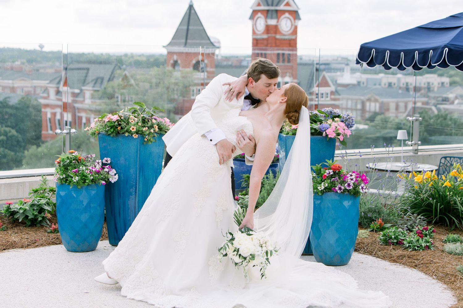 Bride and groom share a romantic dip kiss on a rooftop wedding venue with blue planters and city skyline backdrop.