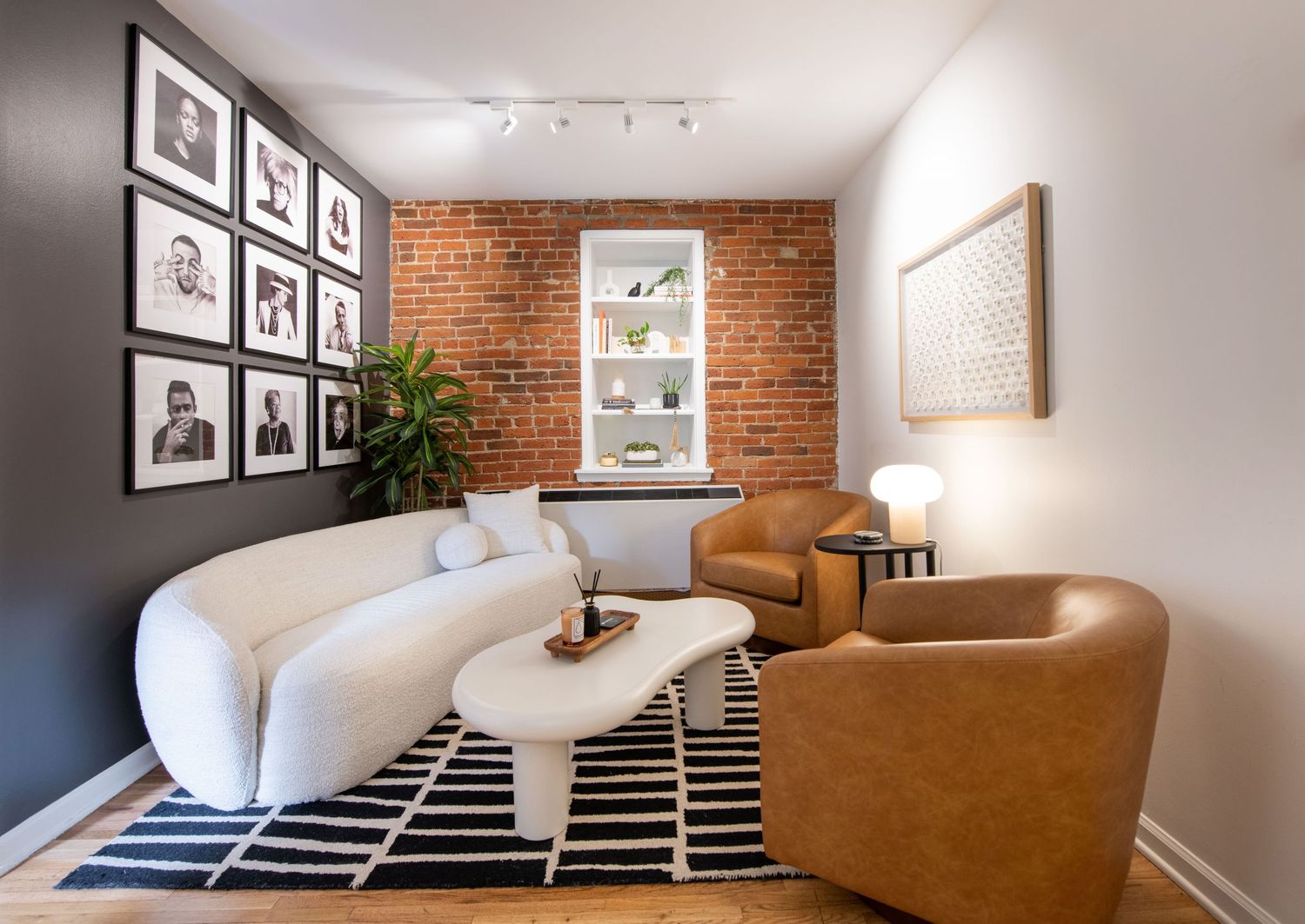 Modern living room with white curved sofa, brown chairs, and exposed brick wall featuring black and white gallery wall.