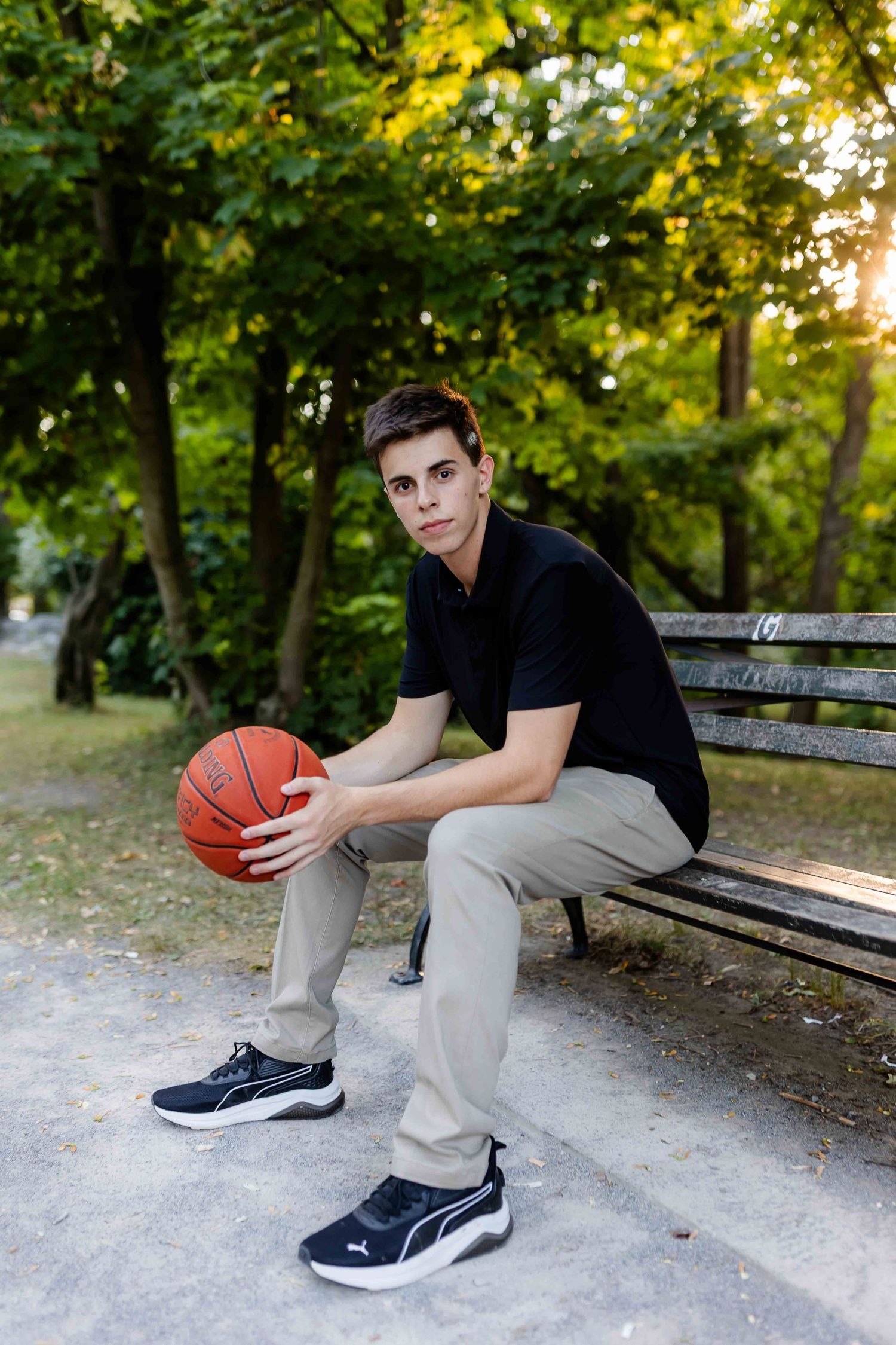 Athletic person in black shirt and khakis sitting on bench with basketball in park setting.