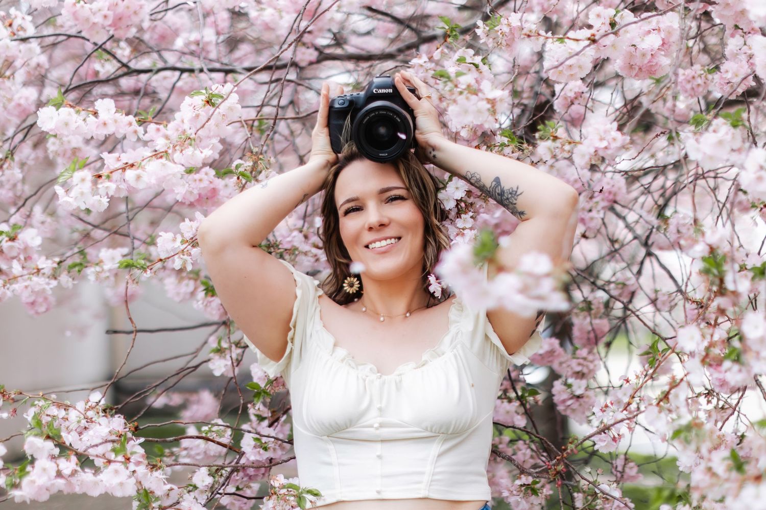 Woman smiling under pink cherry blossom trees holding a camera above her head, wearing a white crop top.