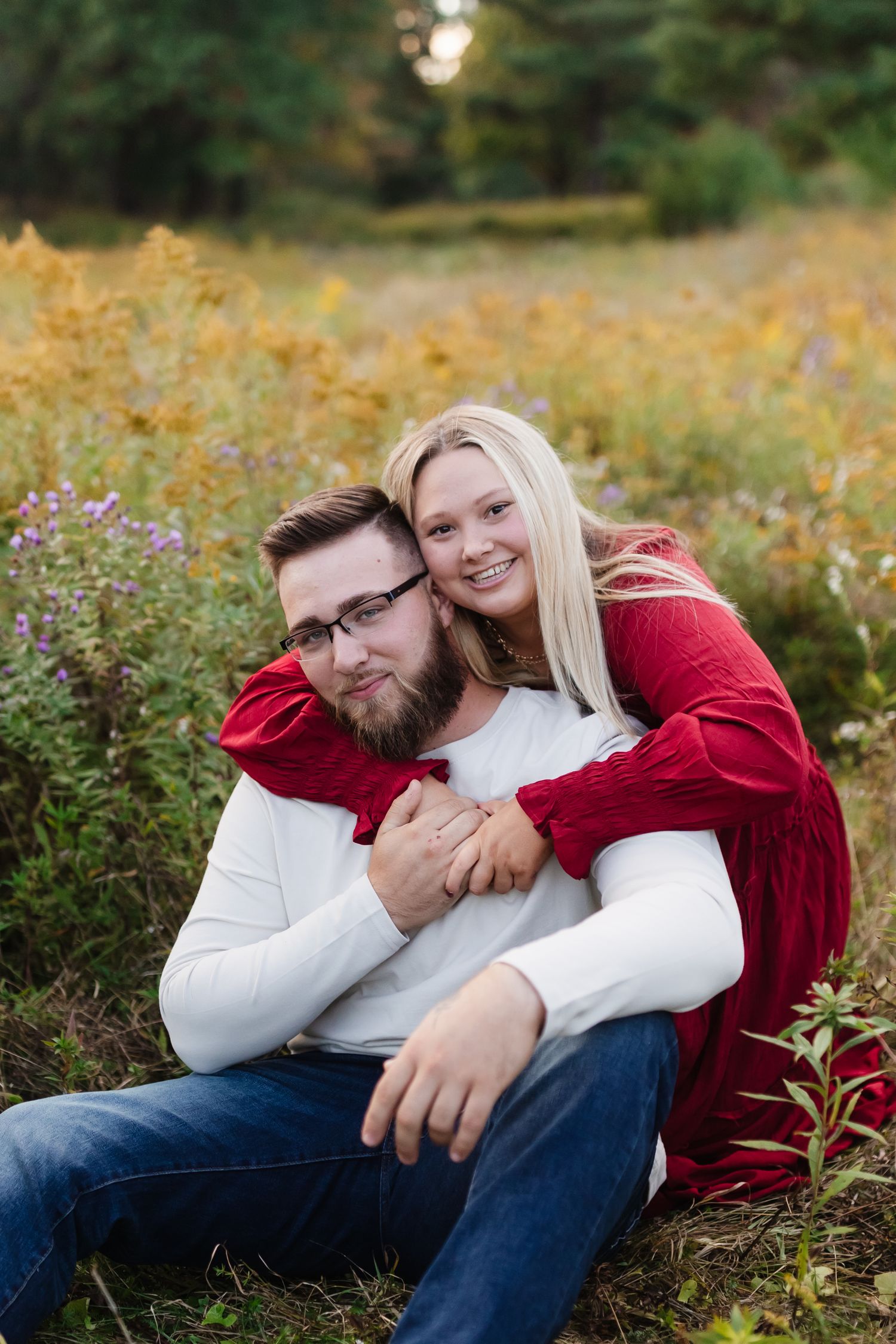A couple wearing matching red and white sweaters embrace while sitting in a wildflower field during autumn.