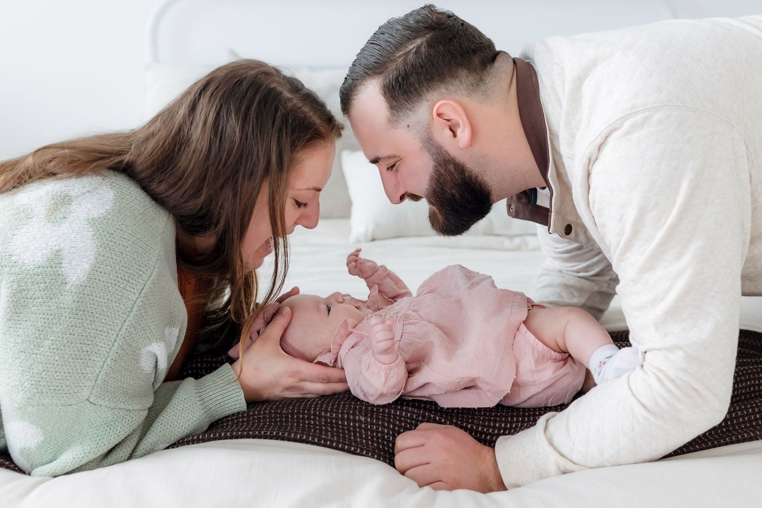 A loving couple leans over their newborn baby lying on a white bed, sharing a tender family moment.