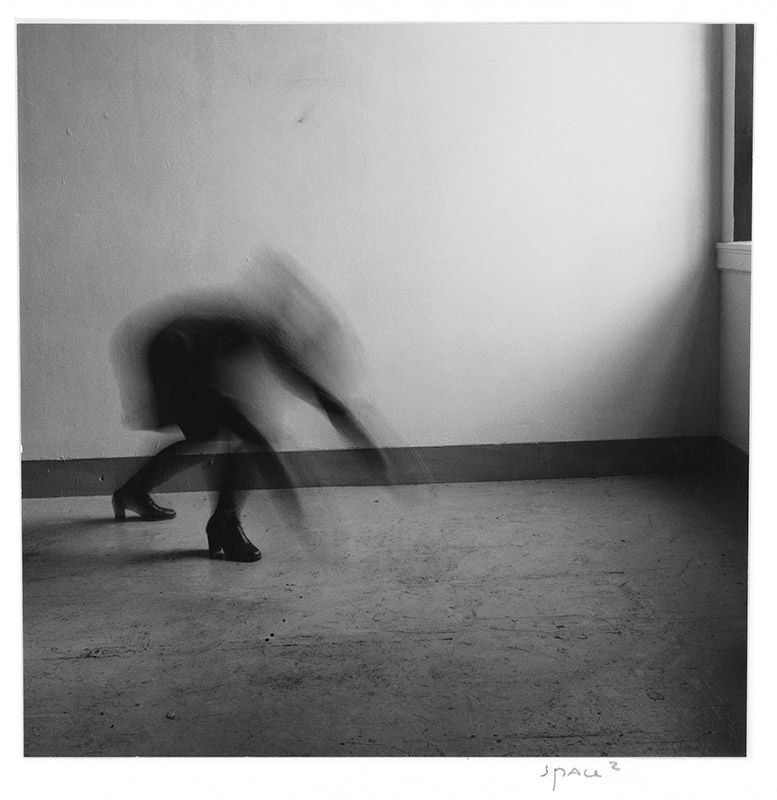 A black and white abstract photograph shows a blurred figure in motion against a stark white wall and dark floor.
