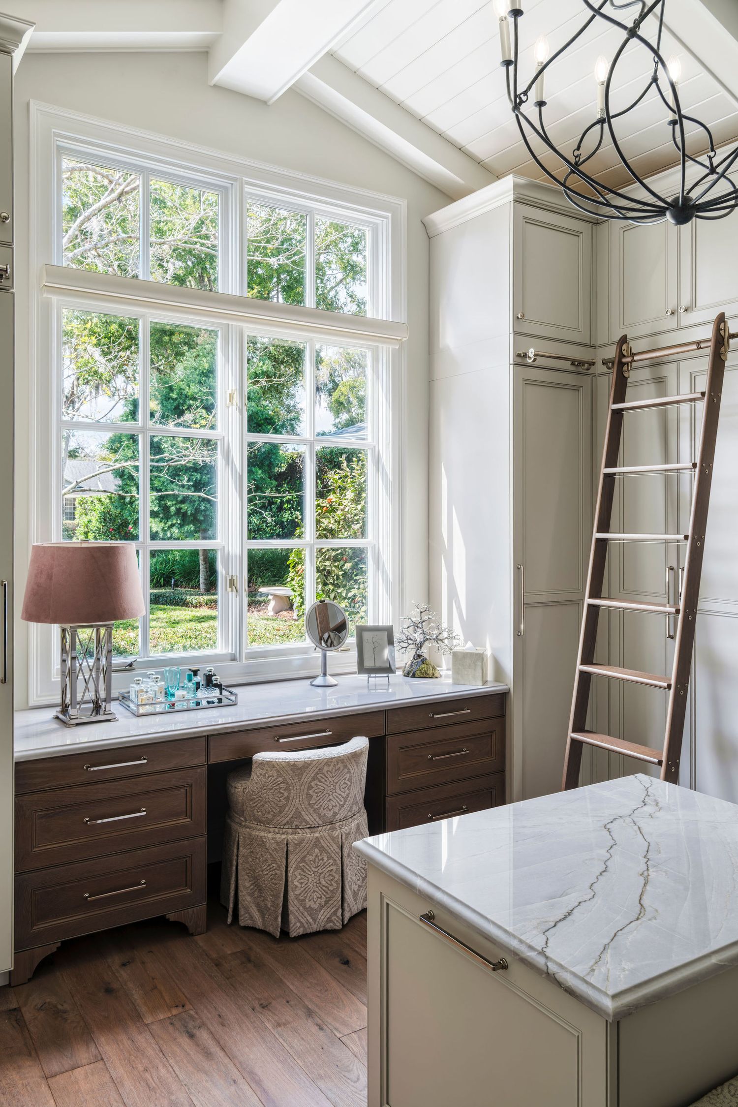 Elegant home office with dark wood built-in desk, marble countertop island, library ladder, and large windows overlooking lush greenery.