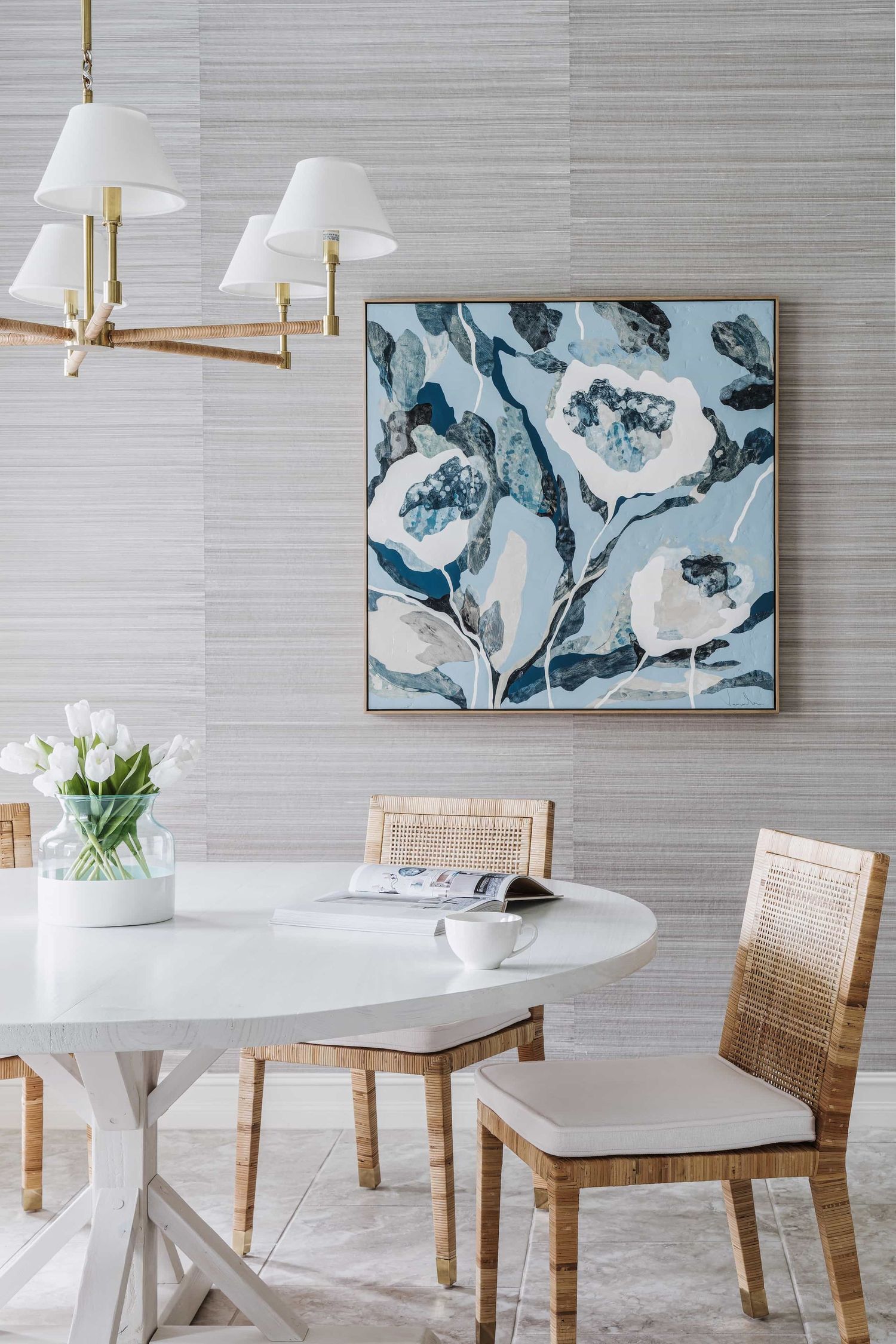 Modern dining space with white round table, rattan chairs, and blue floral artwork on textured gray walls.