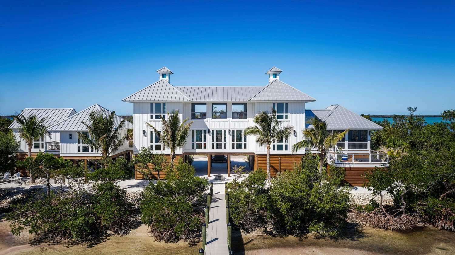 Luxury beachfront white two-story home with metal roof, palm trees, and ocean views under a clear blue sky.
