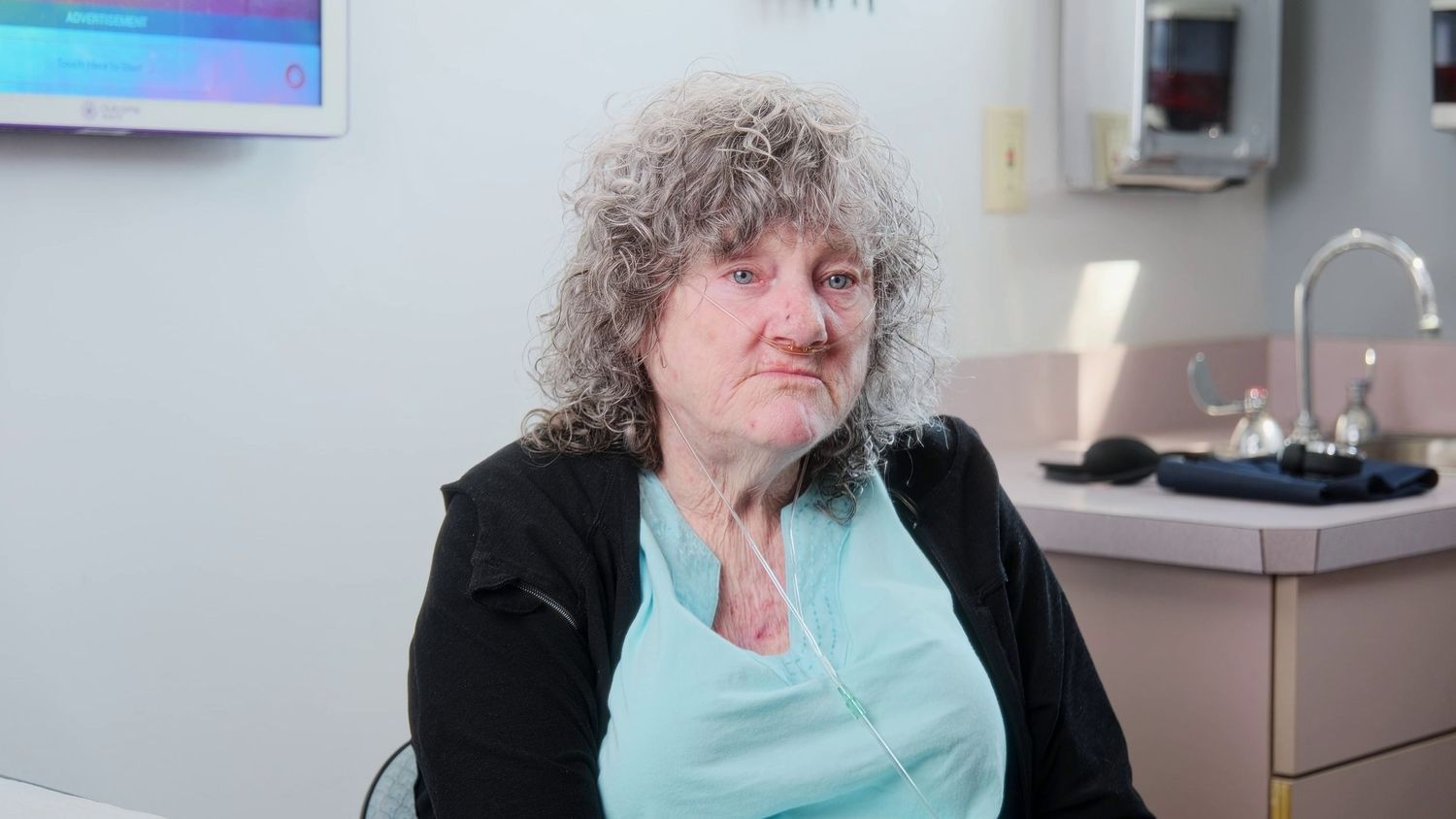 Person in black jacket and turquoise shirt sitting near medical equipment in clinical setting.