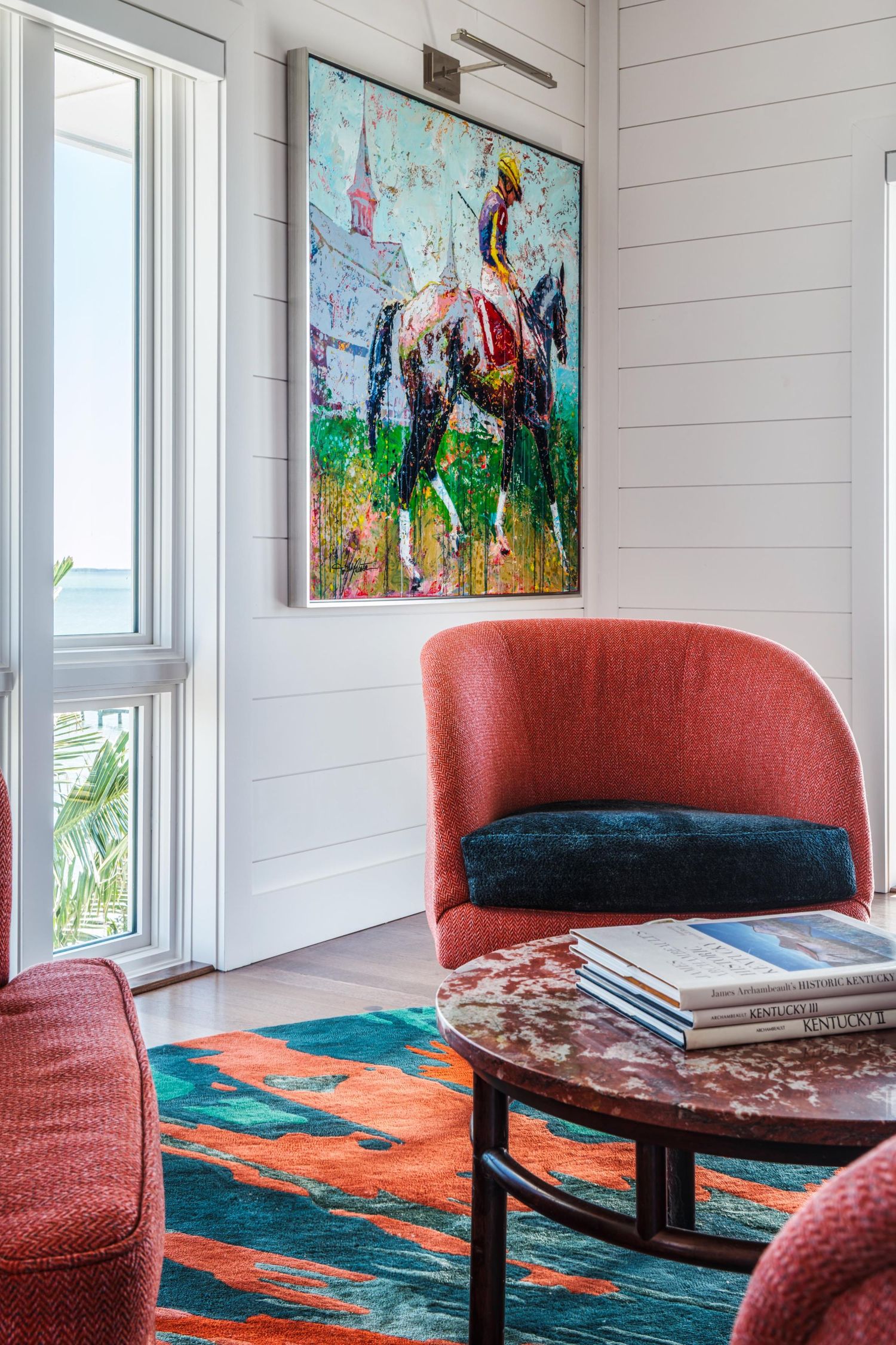 Modern coastal living room featuring coral chairs, vibrant equestrian artwork, and a teal-orange area rug beside a bright window.