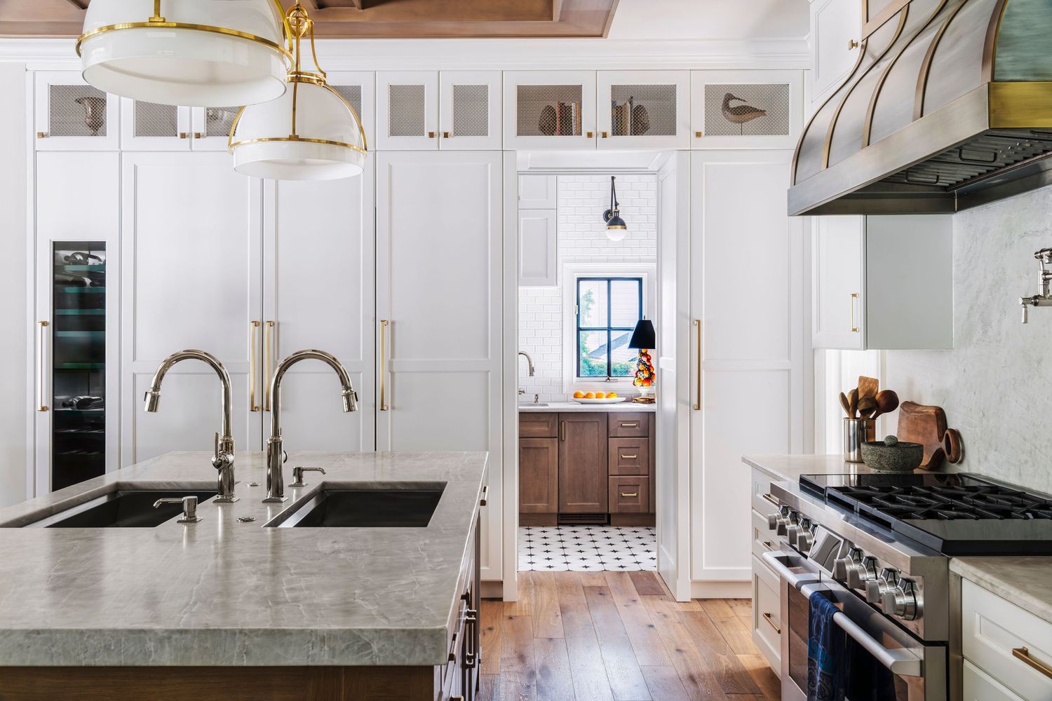 Elegant white kitchen with marble island, brass fixtures, gas range, and hardwood floors leading to a butler's pantry.