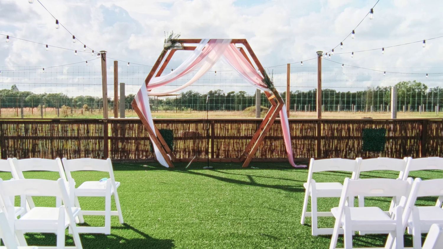 Modern hexagonal wedding arch with pink draping set up on grass lawn with white chairs for outdoor ceremony.