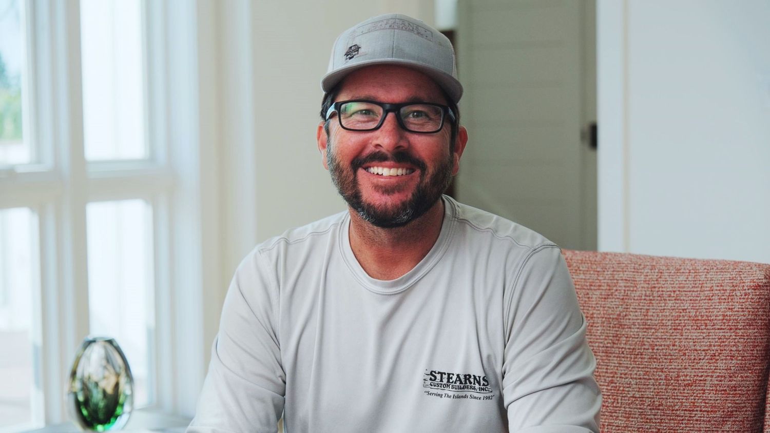 A smiling person wearing glasses and a gray baseball cap sits on a couch in a bright room.