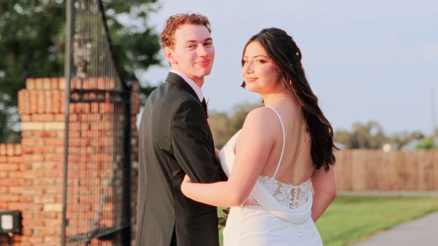 Formal couple posing together against brick wall background in wedding attire.