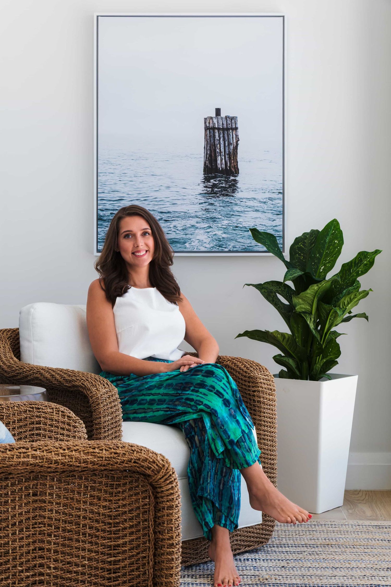 Woman in white top and teal pants sitting in wicker chair beside plant, ocean photo artwork on wall behind her.