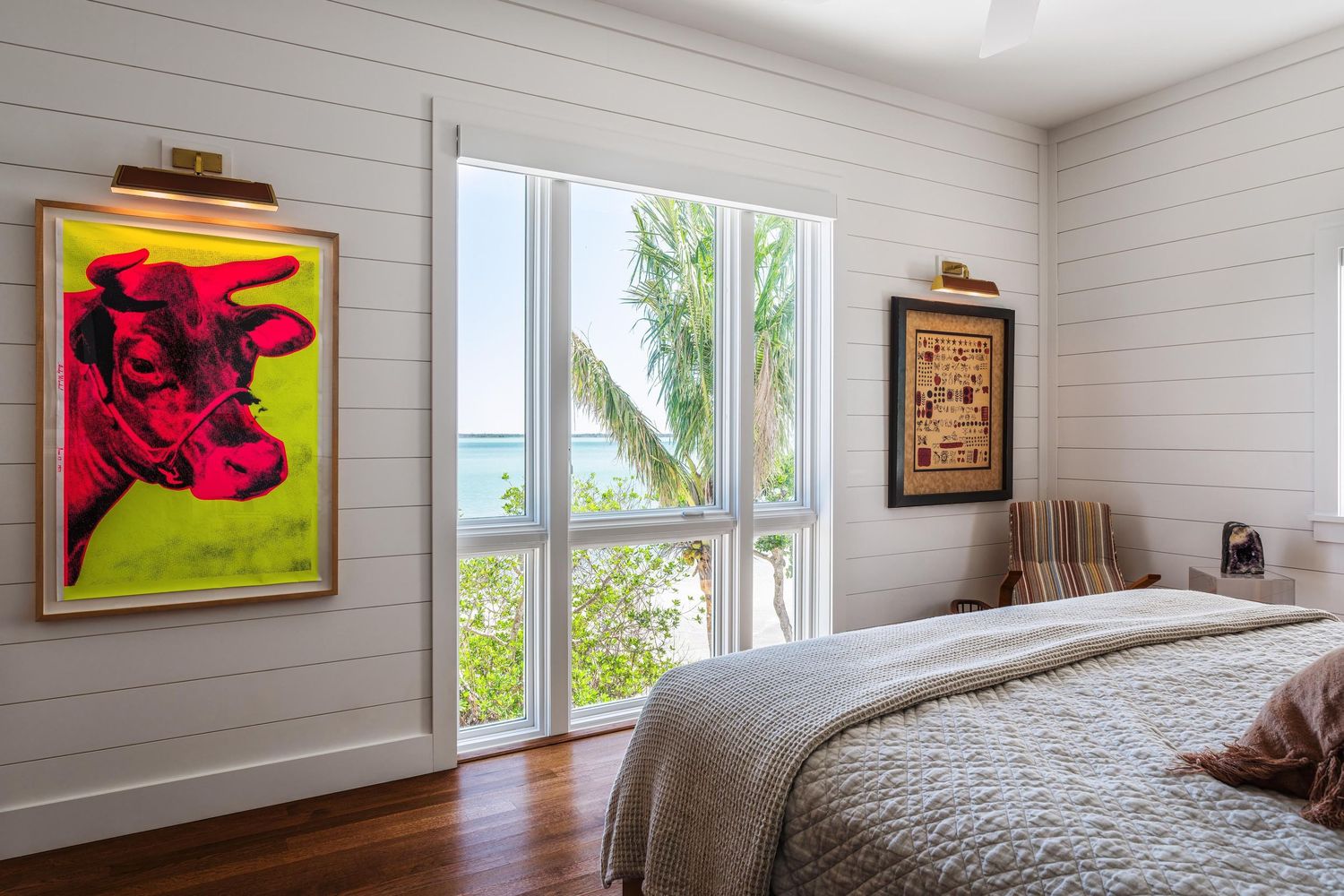 Modern bedroom with bright red artwork, white walls, hardwood floors and tall windows overlooking tropical palm trees.
