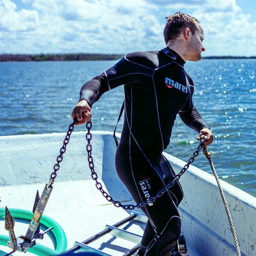 A scuba diver in a black wetsuit handling metal chains on a boat with sparkling blue water in the background for sea restoration efforts.