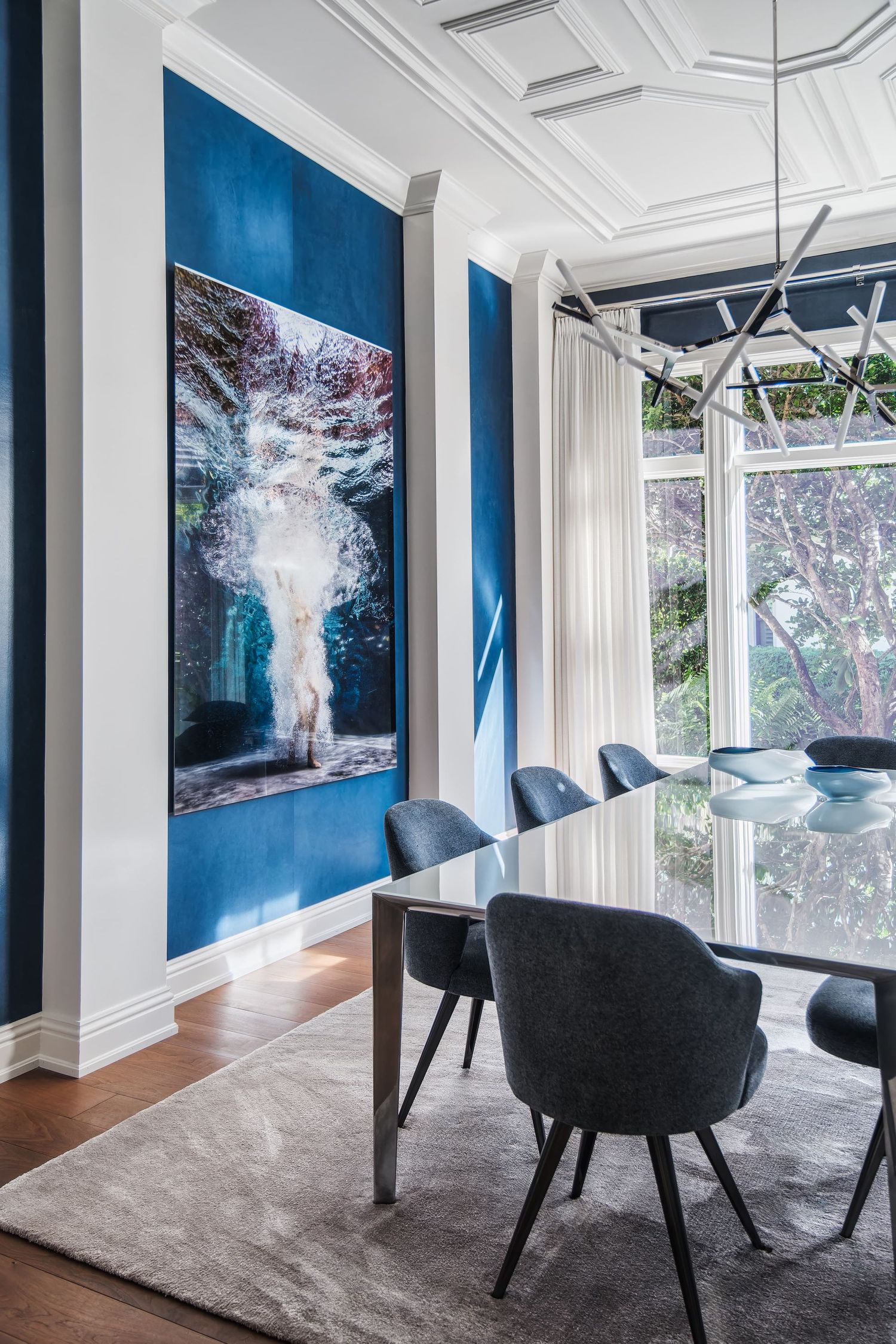 Modern dining room with blue accent walls, waterfall artwork, and black upholstered chairs around glass table.