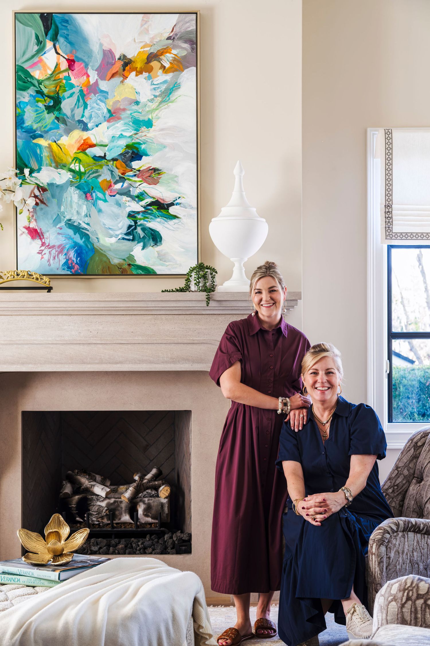 Two women pose in an elegant living room beside a fireplace with colorful abstract artwork above the mantle.