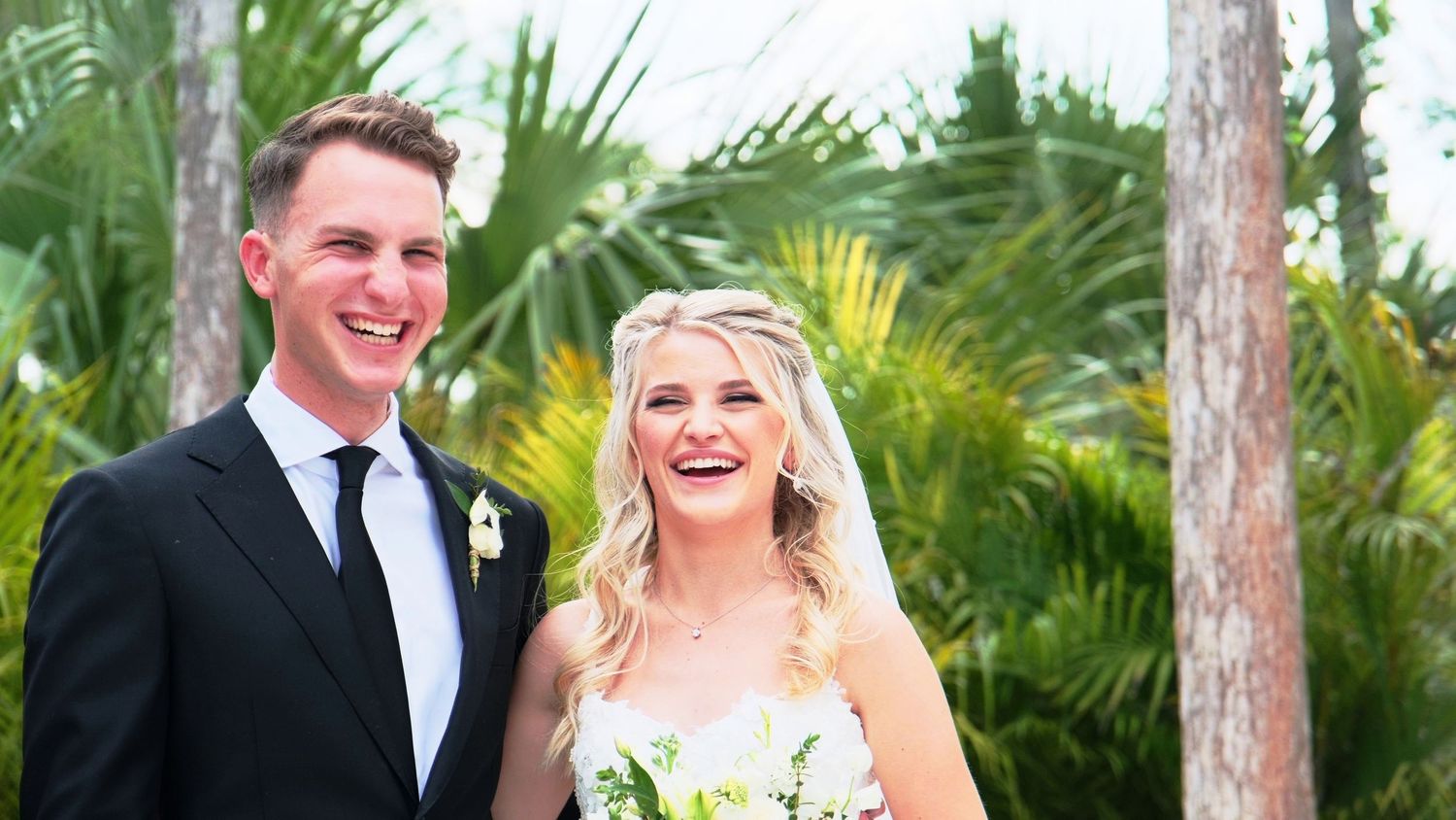 Wedding couple standing together among tropical palm trees in formal attire.