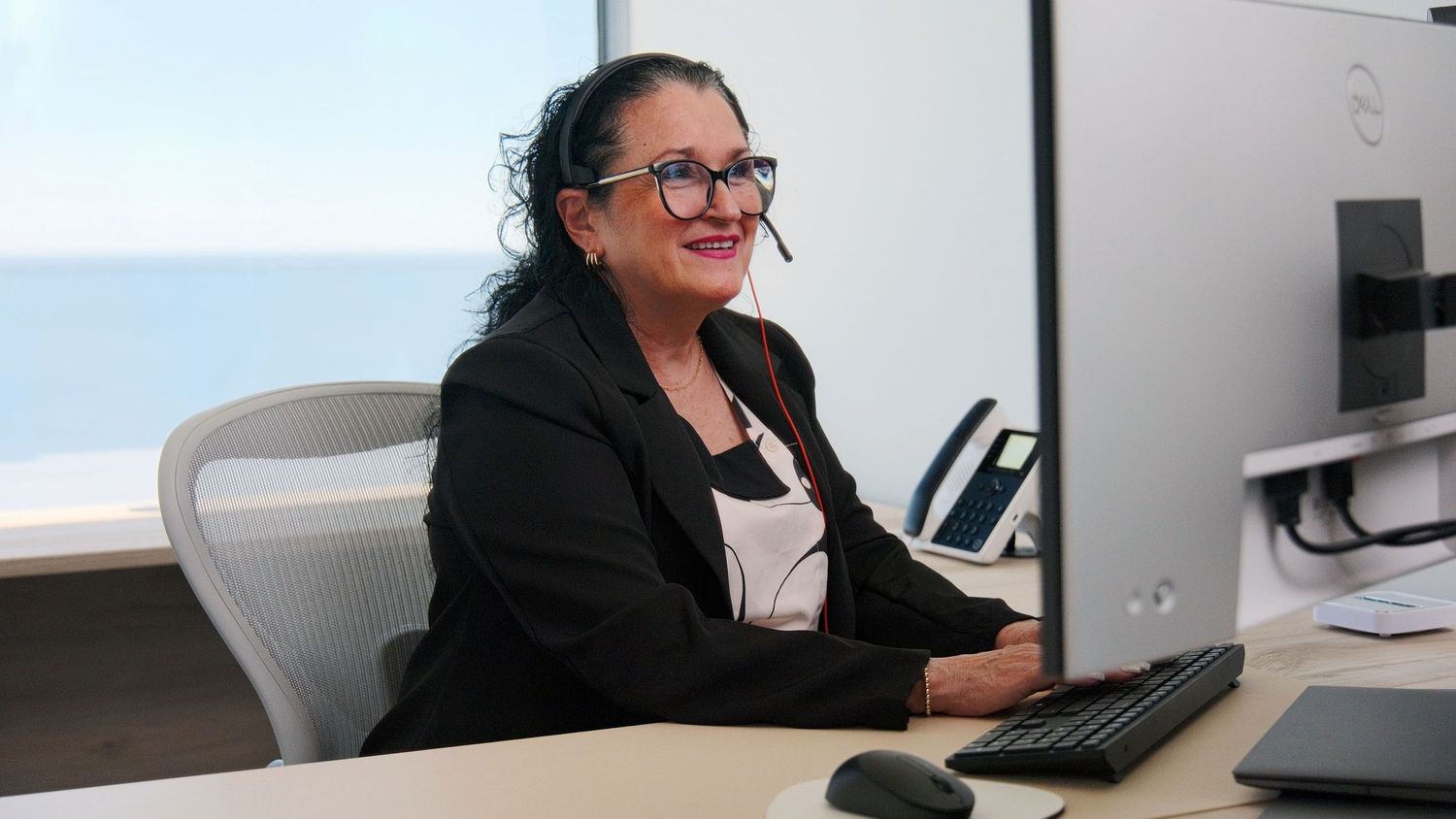 Professional woman with headset smiling at computer desk in modern office with ocean view.