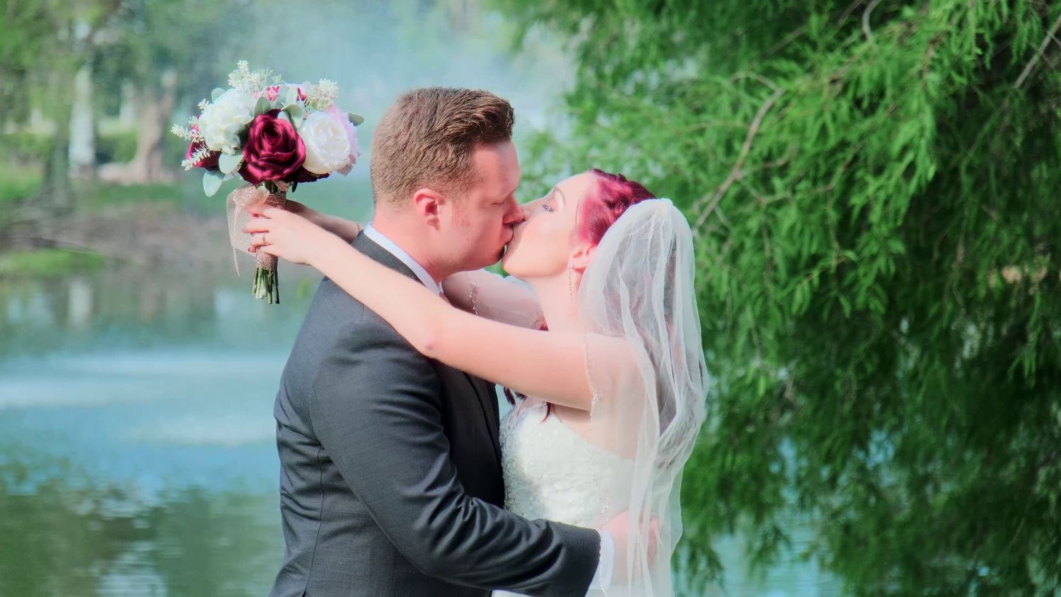 Wedding moment captured with couple embracing near waterside with lush greenery.