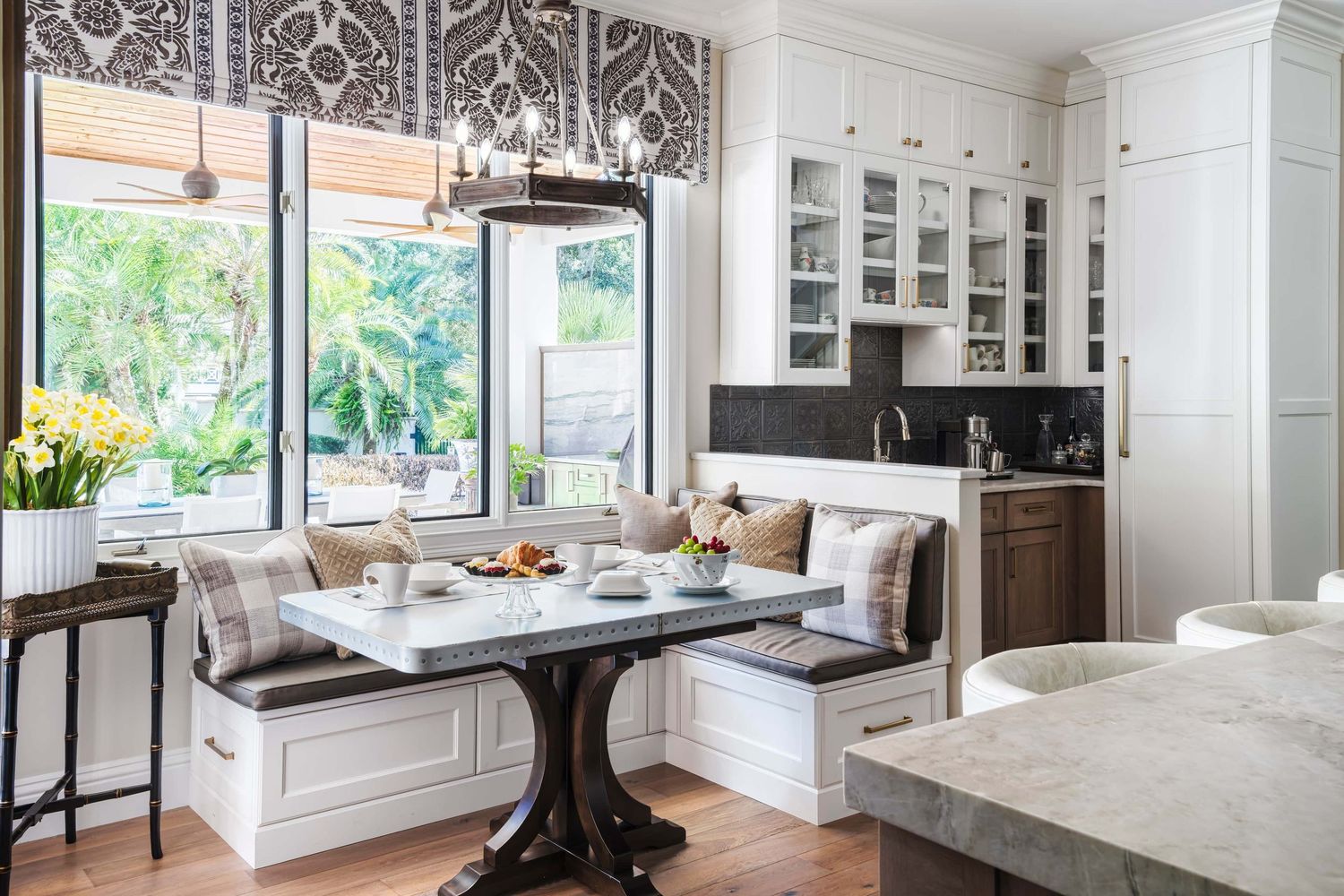 Elegant kitchen breakfast nook with built-in banquette seating, round pedestal table, white cabinetry, and patterned Roman shade.