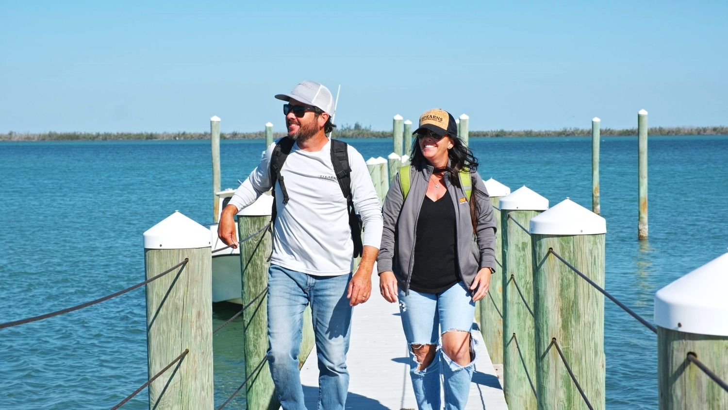 Two people walking on a wooden pier with boats and turquoise blue water on a sunny day.