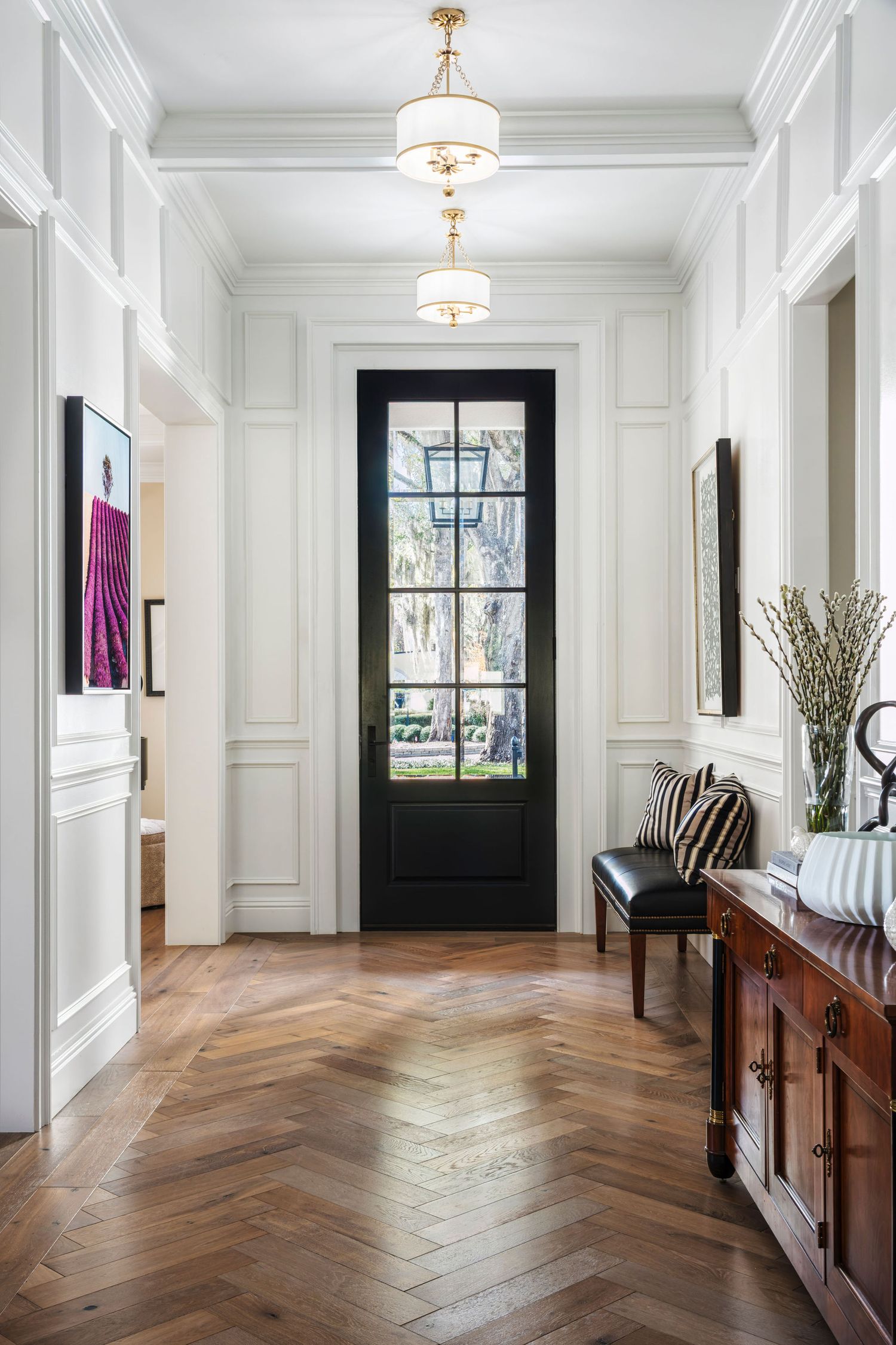 Elegant foyer with herringbone wood floors, white wainscoting, black glass front door, pendant lights, and a wooden console table.