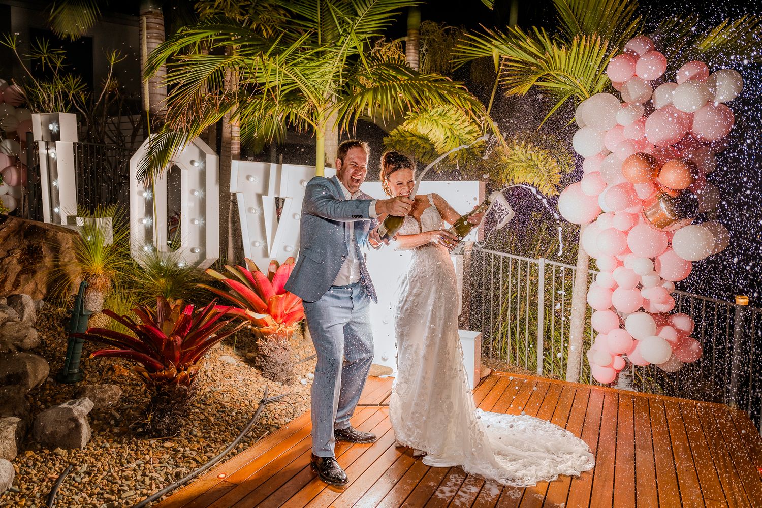 A romantic wedding couple dances on a deck decorated with pink balloon garlands and tropical palm trees.