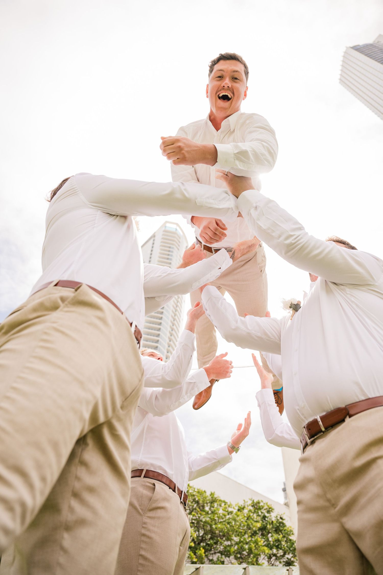 Groomsmen in khaki pants toss someone up in celebration against a bright urban backdrop.