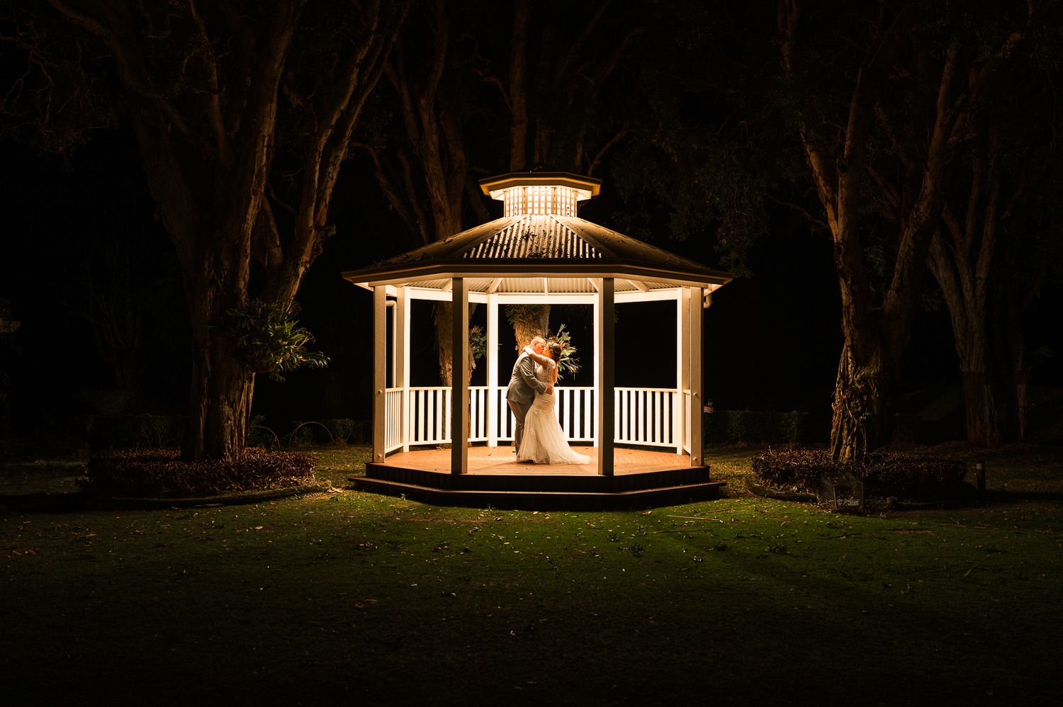 An illuminated white gazebo glows against the dark night sky during a romantic wedding moment.