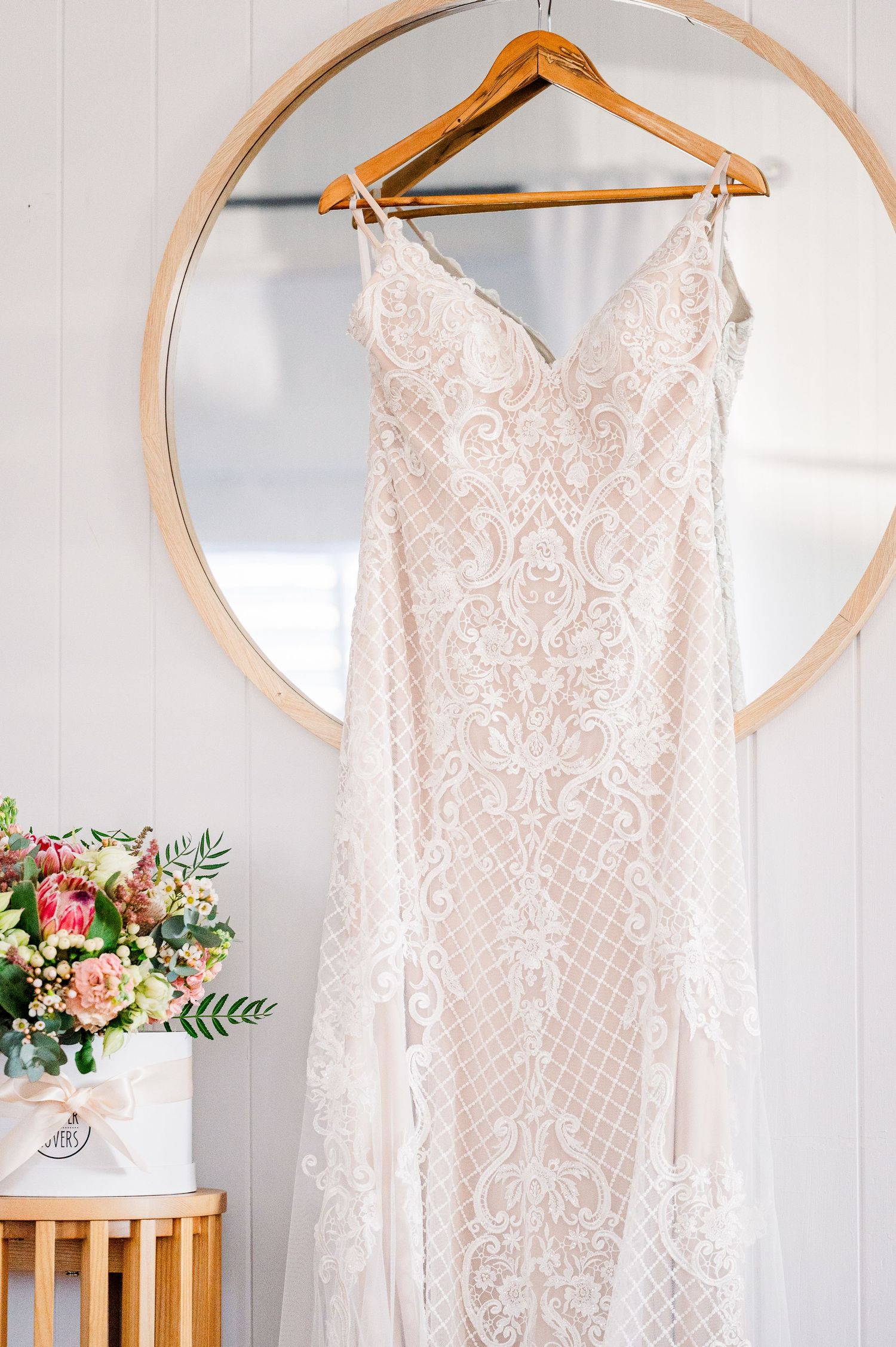 Elegant lace wedding dress hanging on wooden hanger against round mirror with floral arrangement.