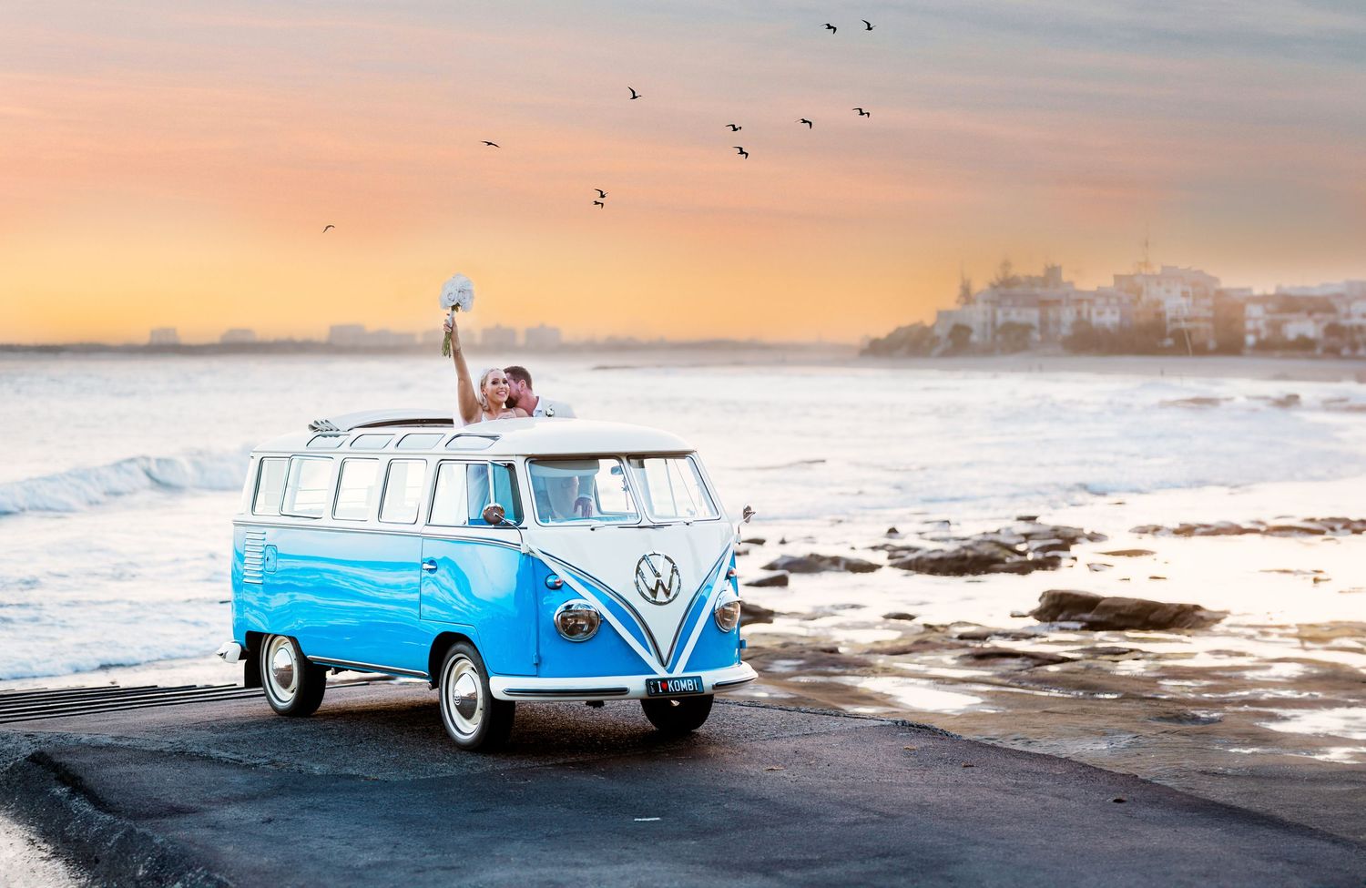 Classic blue Volkswagen van parked by the beach at sunset with birds flying overhead.