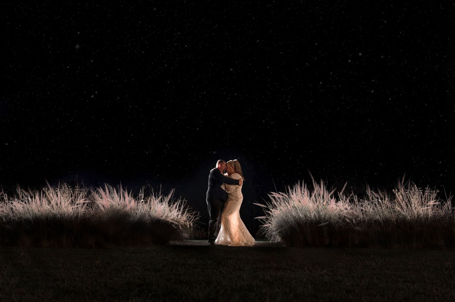 A silhouetted couple sharing an intimate moment under dramatic lighting with illuminated grass in the dark background.