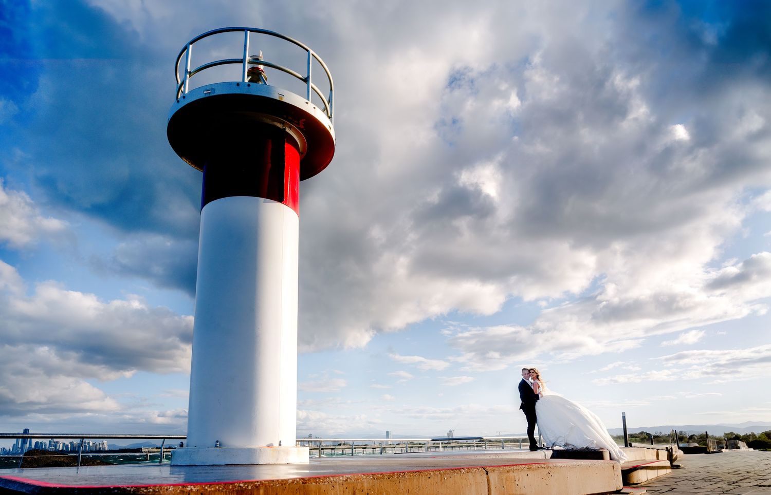 White lighthouse with red stripe against dramatic cloudy sky on a waterfront.