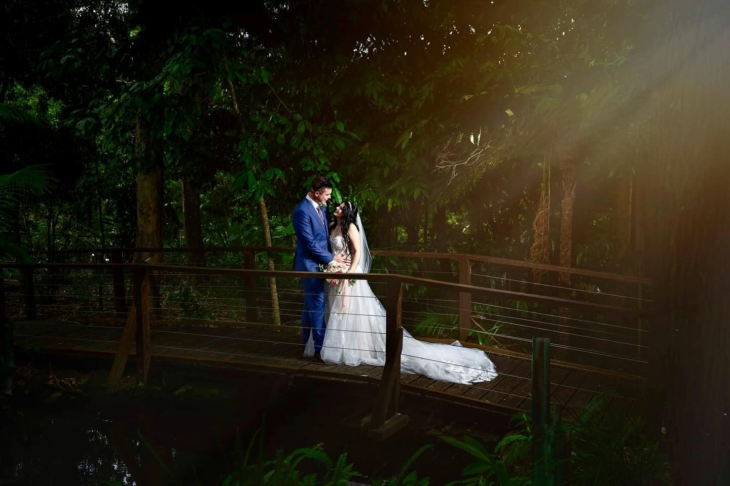 Romantic woodland bridge wedding photo with dramatic sunlight filtering through trees.