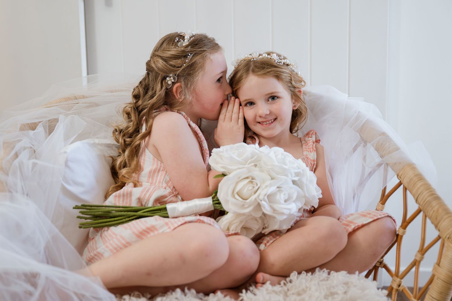 Two flower girls sharing secrets and giggles while holding a white bouquet.