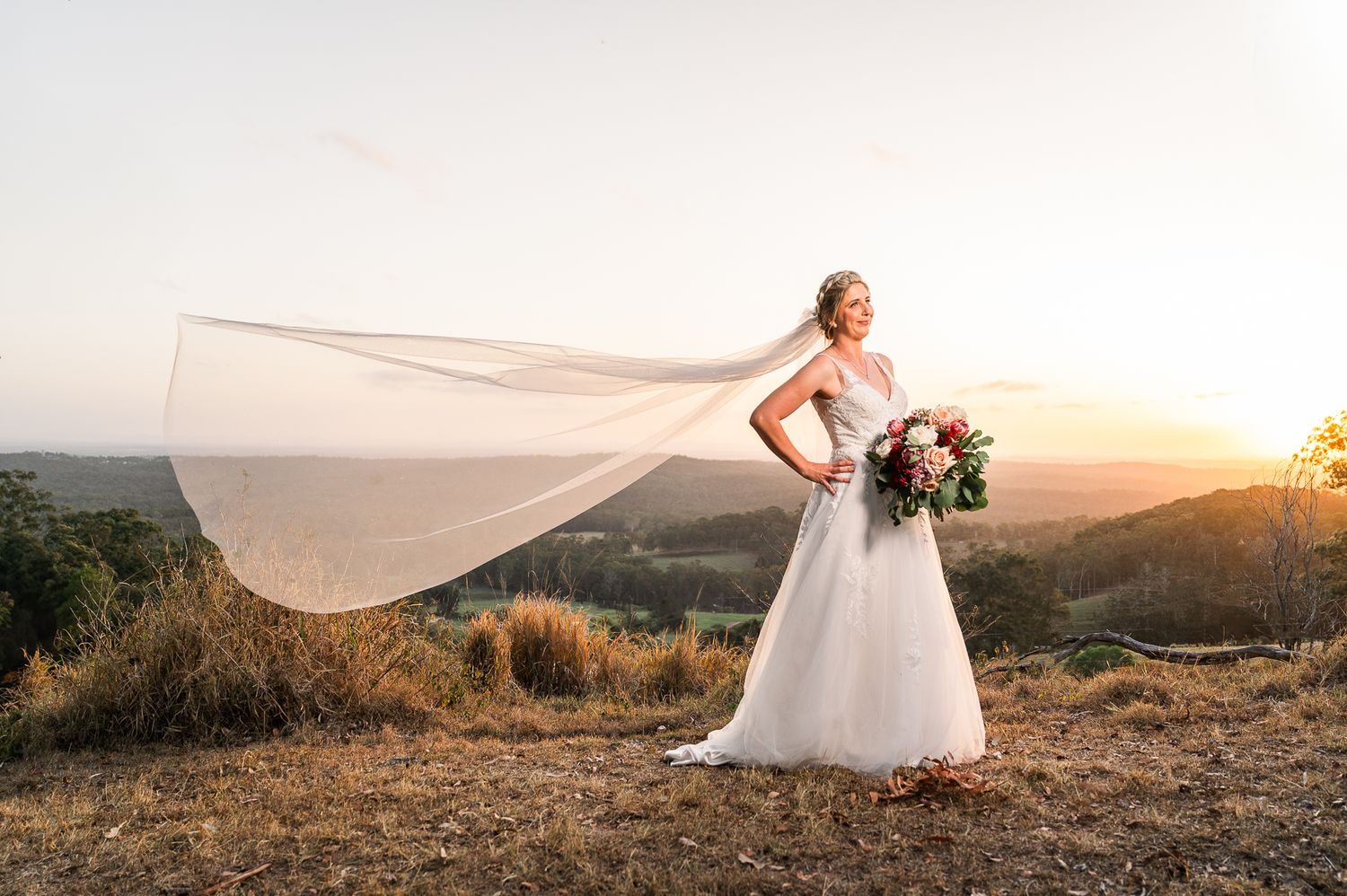 A flowing white wedding dress and veil catch the golden sunset light on a hilltop.