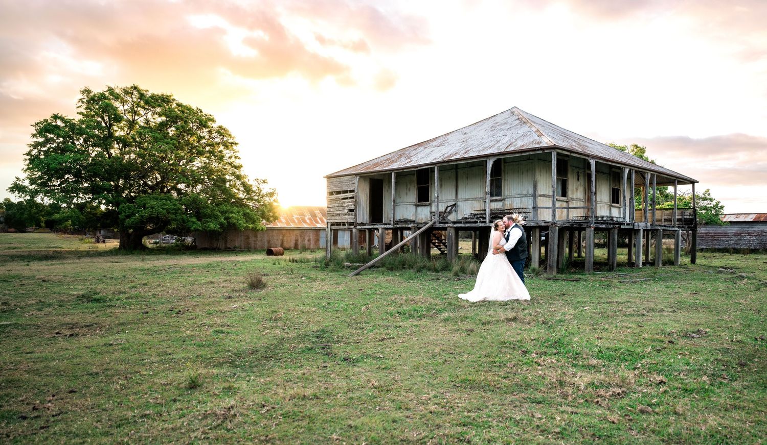 Rustic wedding scene with vintage farmhouse and large tree at sunset.