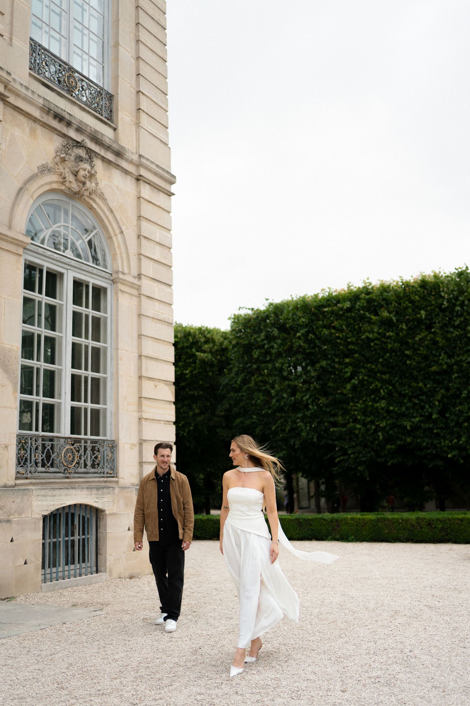 A couple walks playfully and romantically along a gravel path outside an elegant French chateau with tall hedges. Paris film photographer.