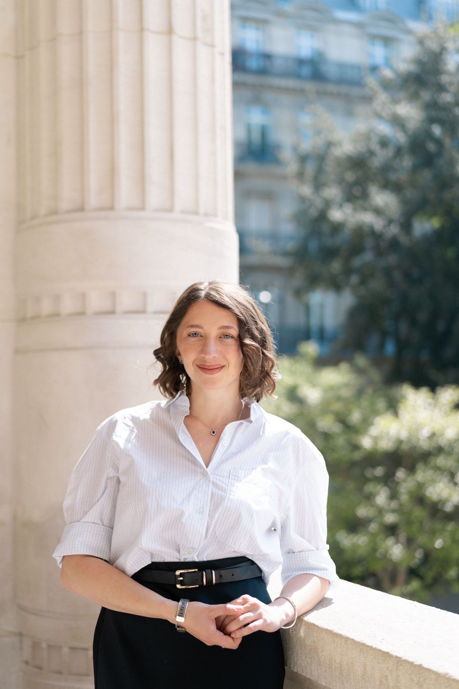 Professional portrait of someone in a crisp white button-down shirt and black skirt standing next to a classical column outdoors.