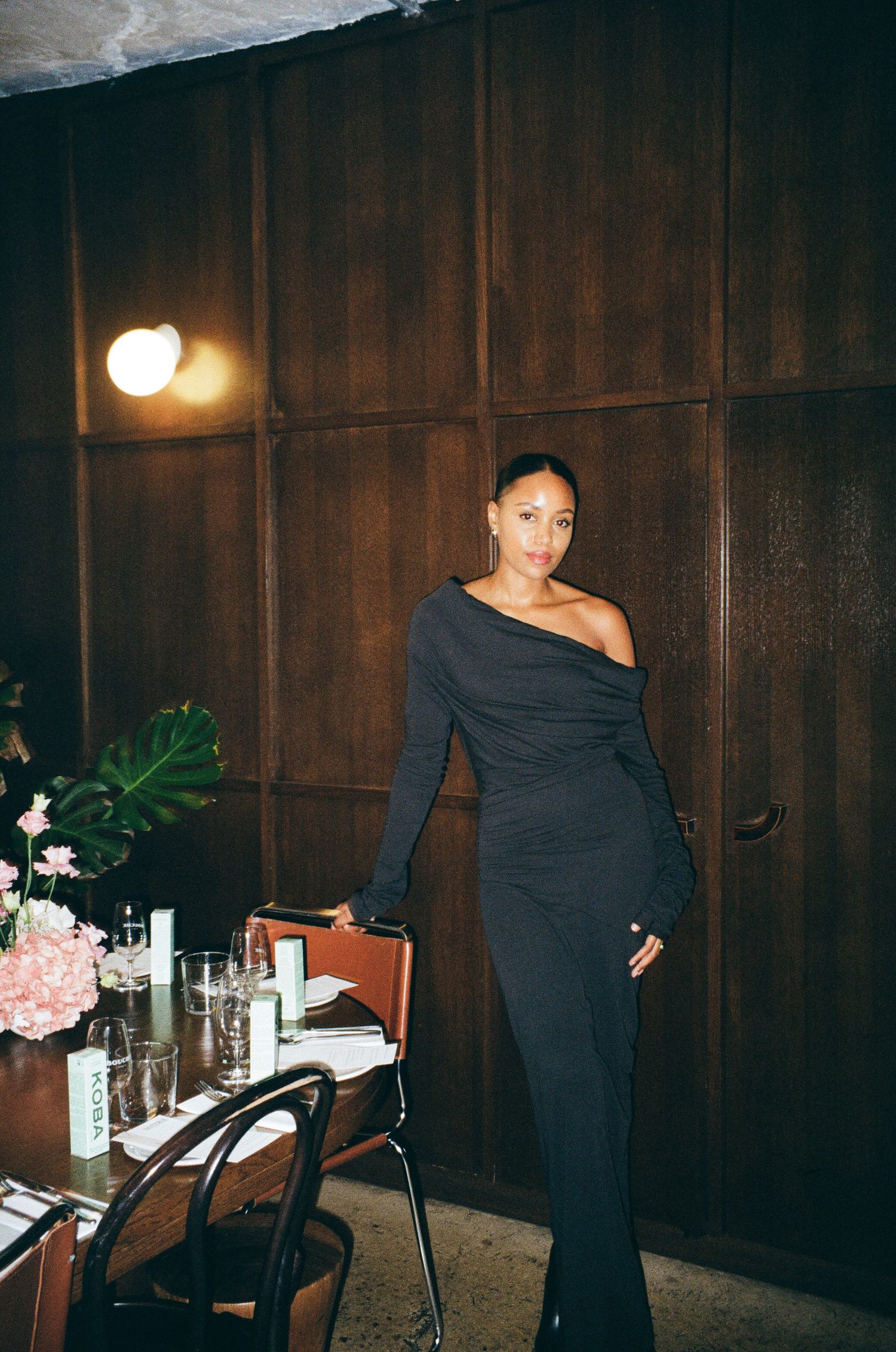 A figure in a black dress poses gracefully against wood-paneled walls in a dimly lit interior space.