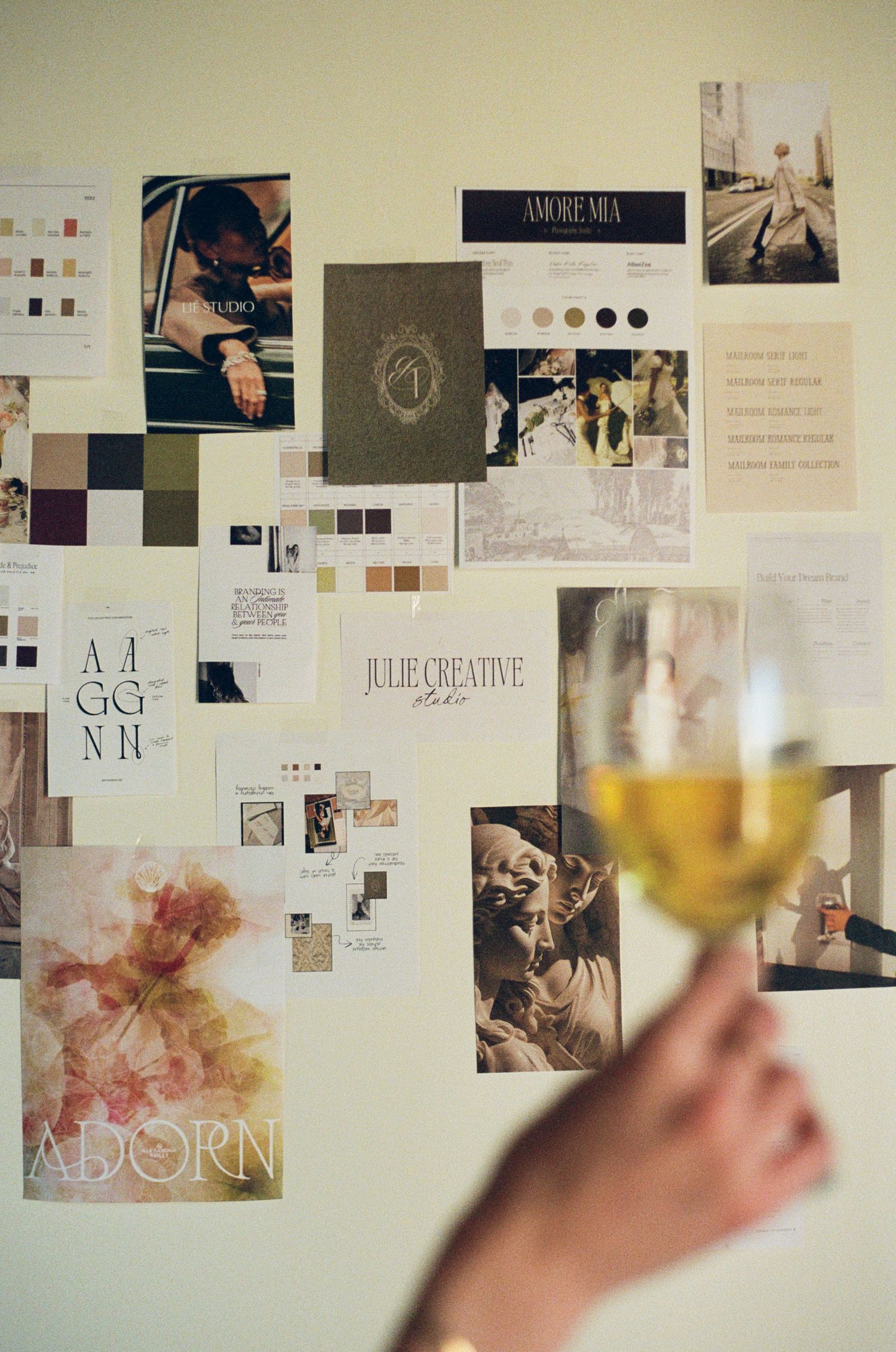 A mood board with photos and design elements on a wall, with someone holding a glass of white wine in the foreground.