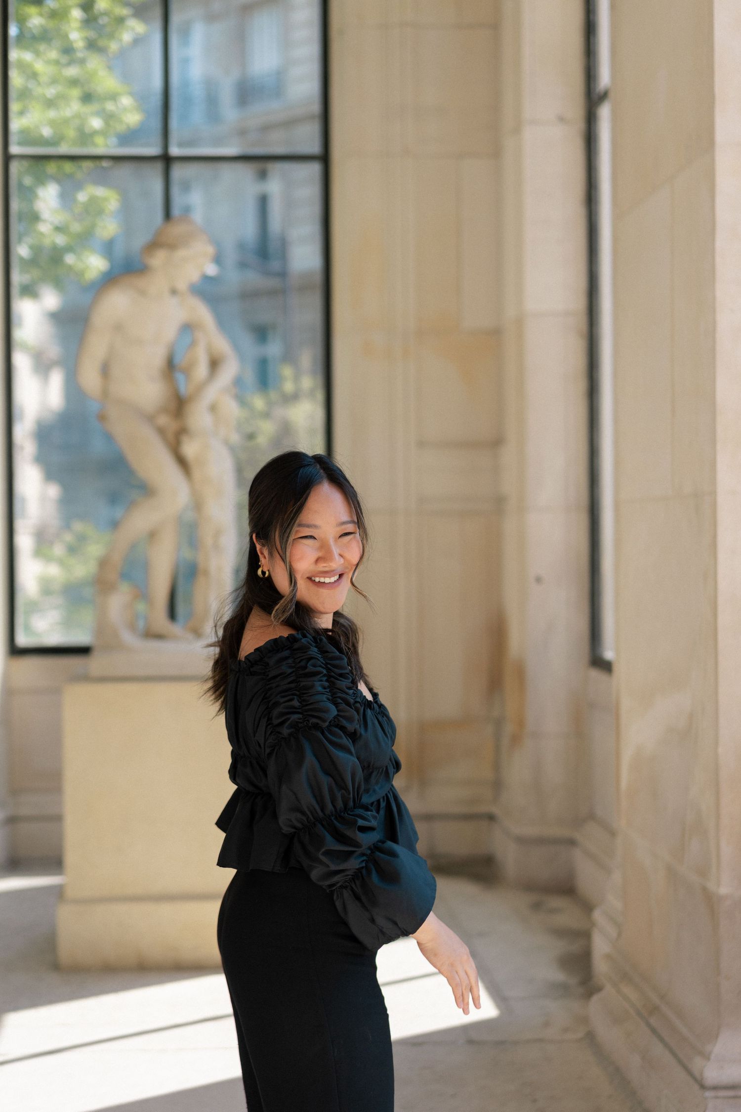 Person wearing a black dress with ruffled shoulder detail poses joyfully in a sunlit colonnade hallway.