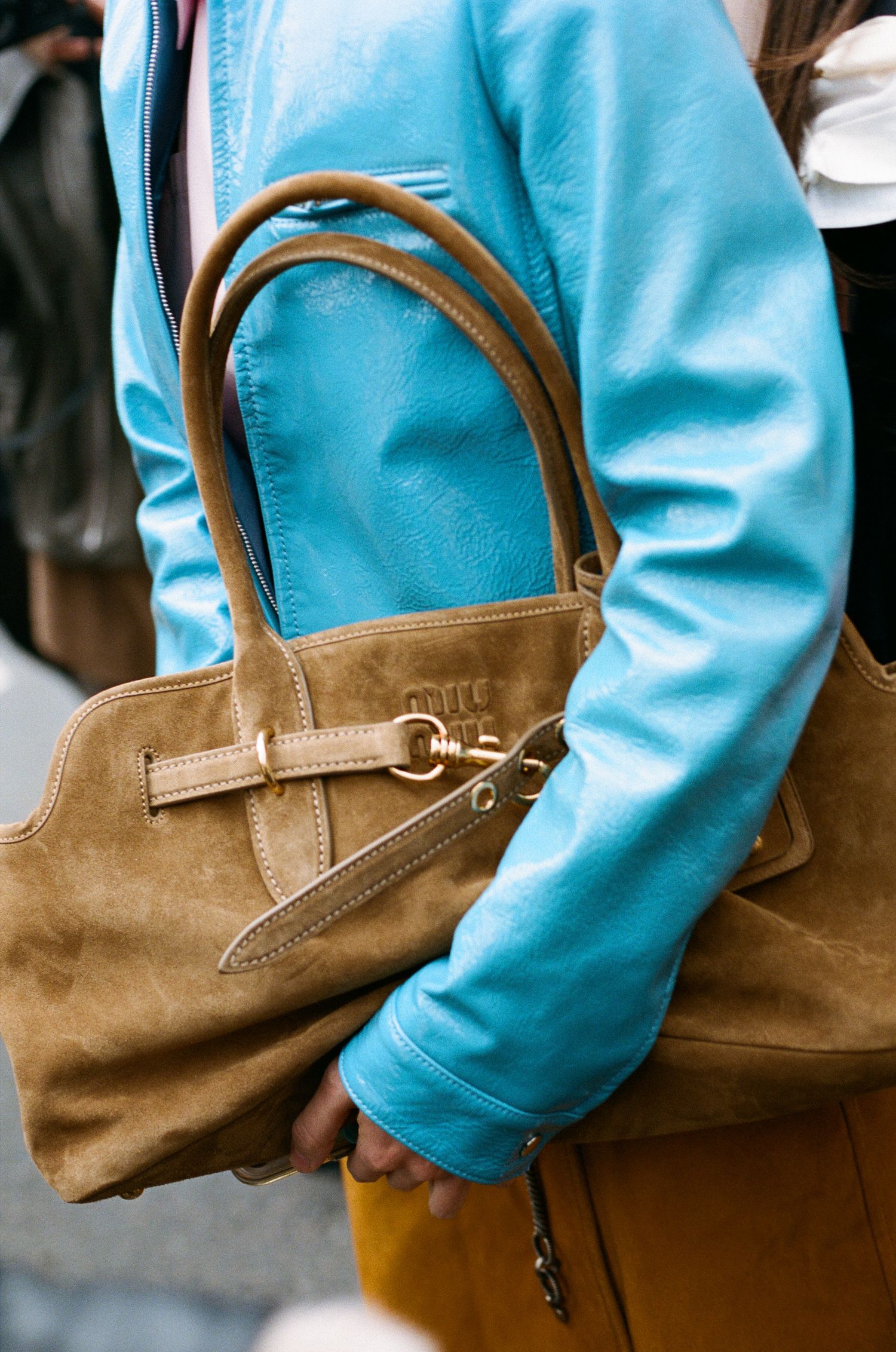 A brown suede handbag with leather handles is held against a light blue fabric background.